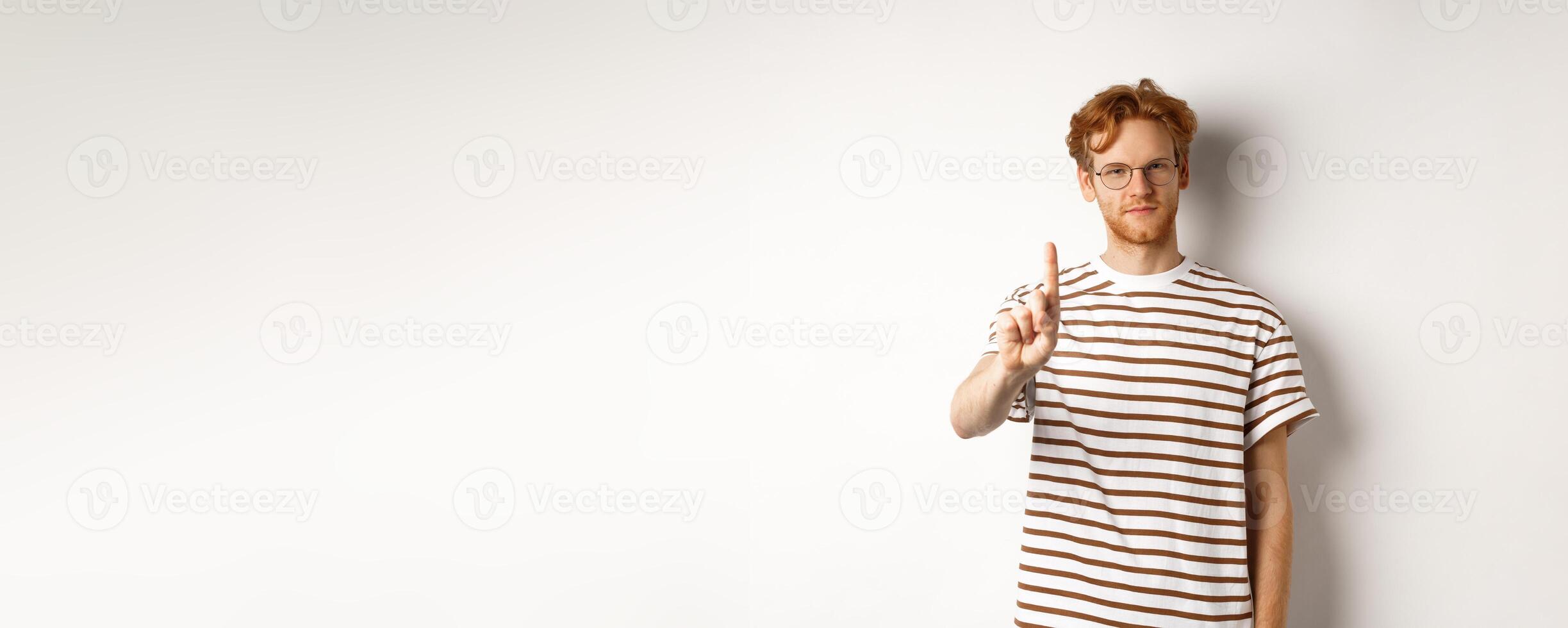 Young man with red hair and glasses shaking finger in prohibition, taboo gesture, disapprove and stop something, standing over white background photo