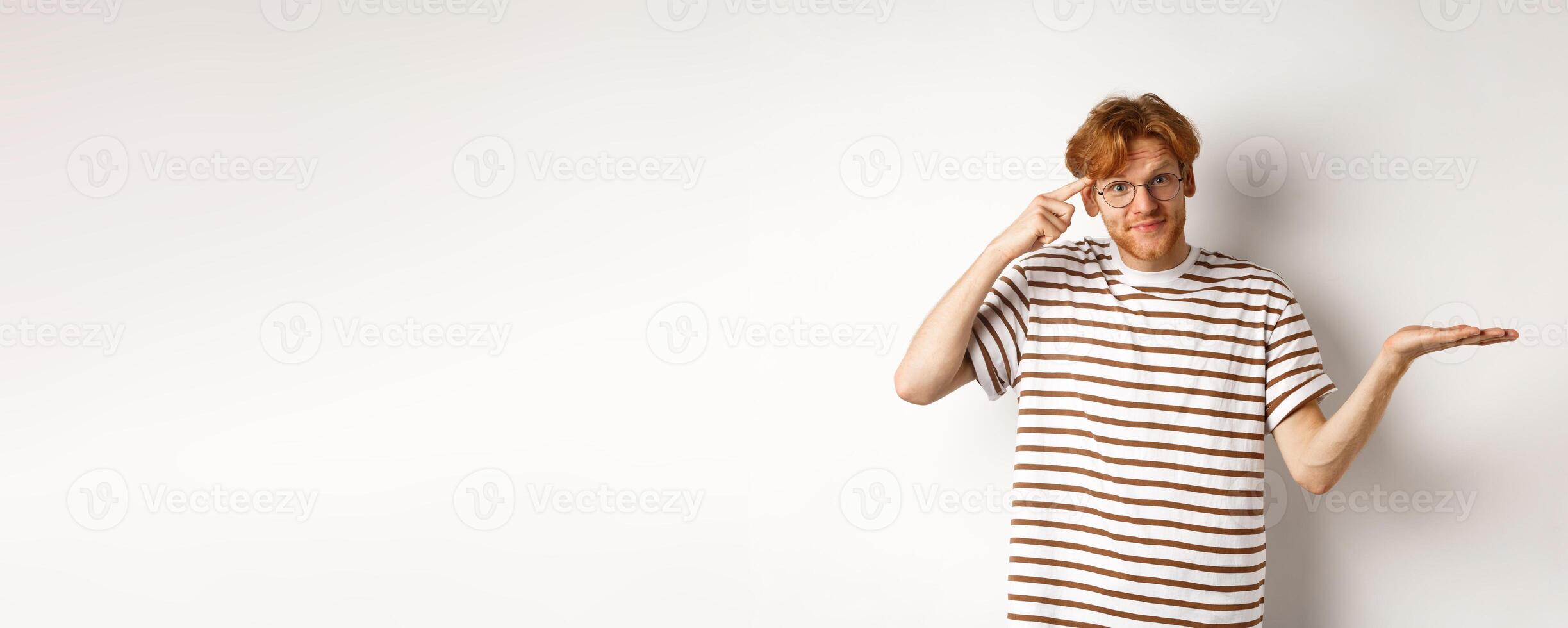 Are you stupid. Confused redhead guy in glasses shrugging, pointing at head and staring skeptical at camera, white background photo