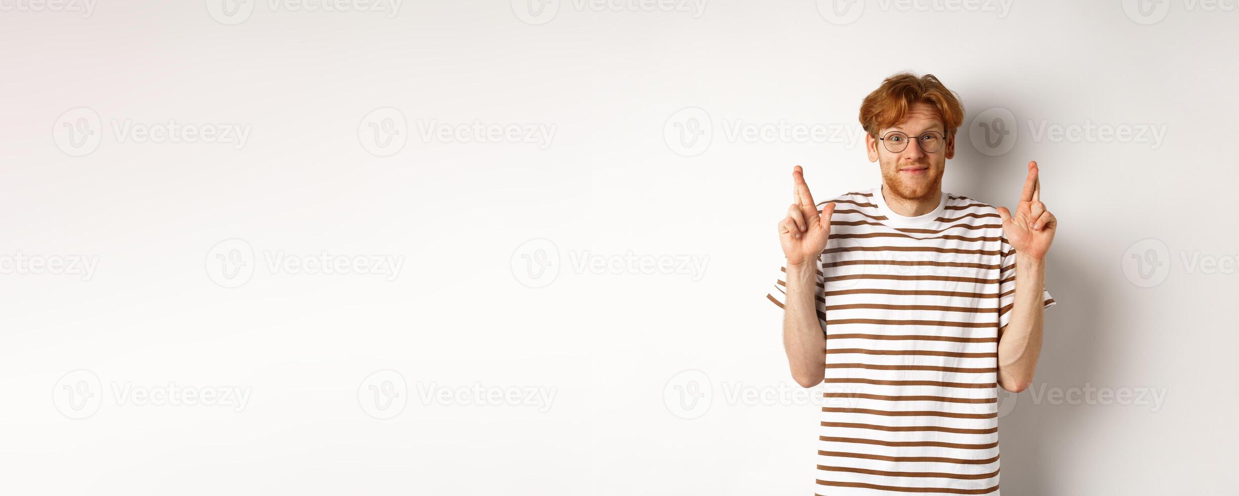 Hopeful young man with red hair and glasses cross fingers for good luck, making wish, standing over white background photo