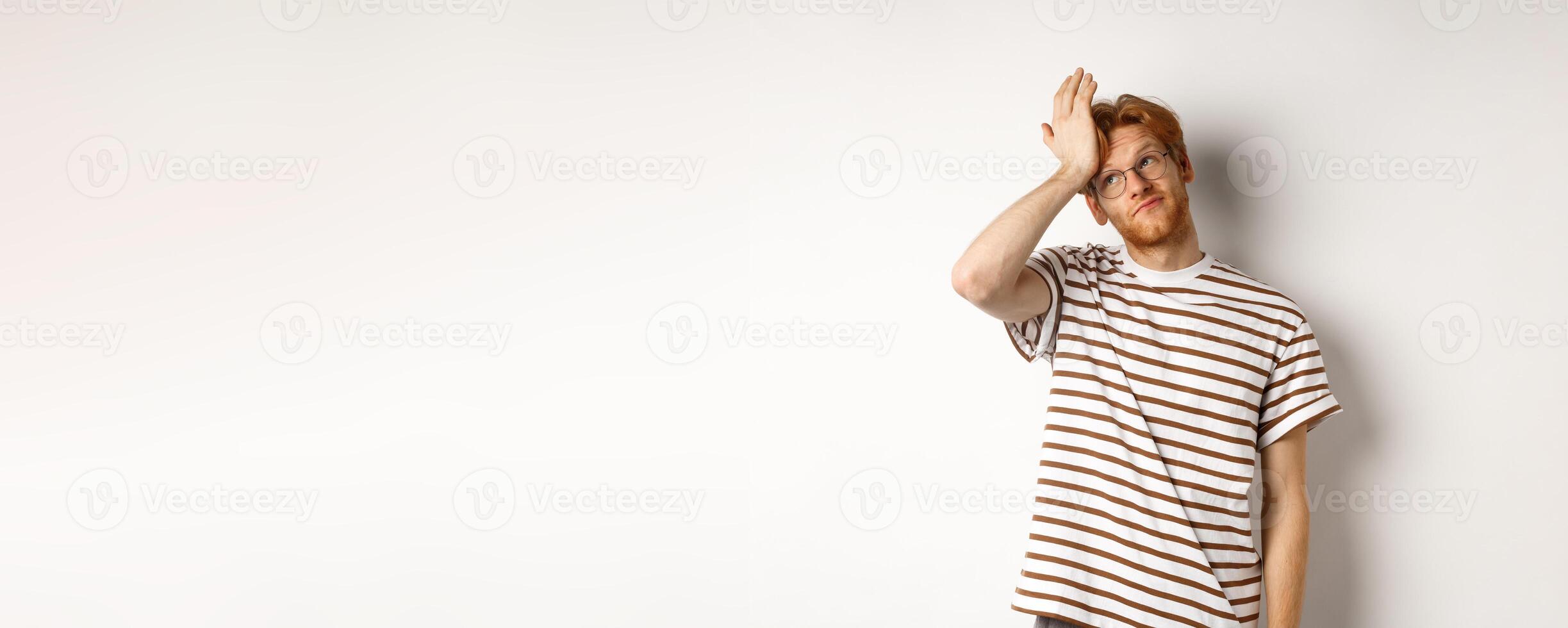 Tired young man with red hair and glasses, roll eyes and making facepalm bothered, standing over white background photo