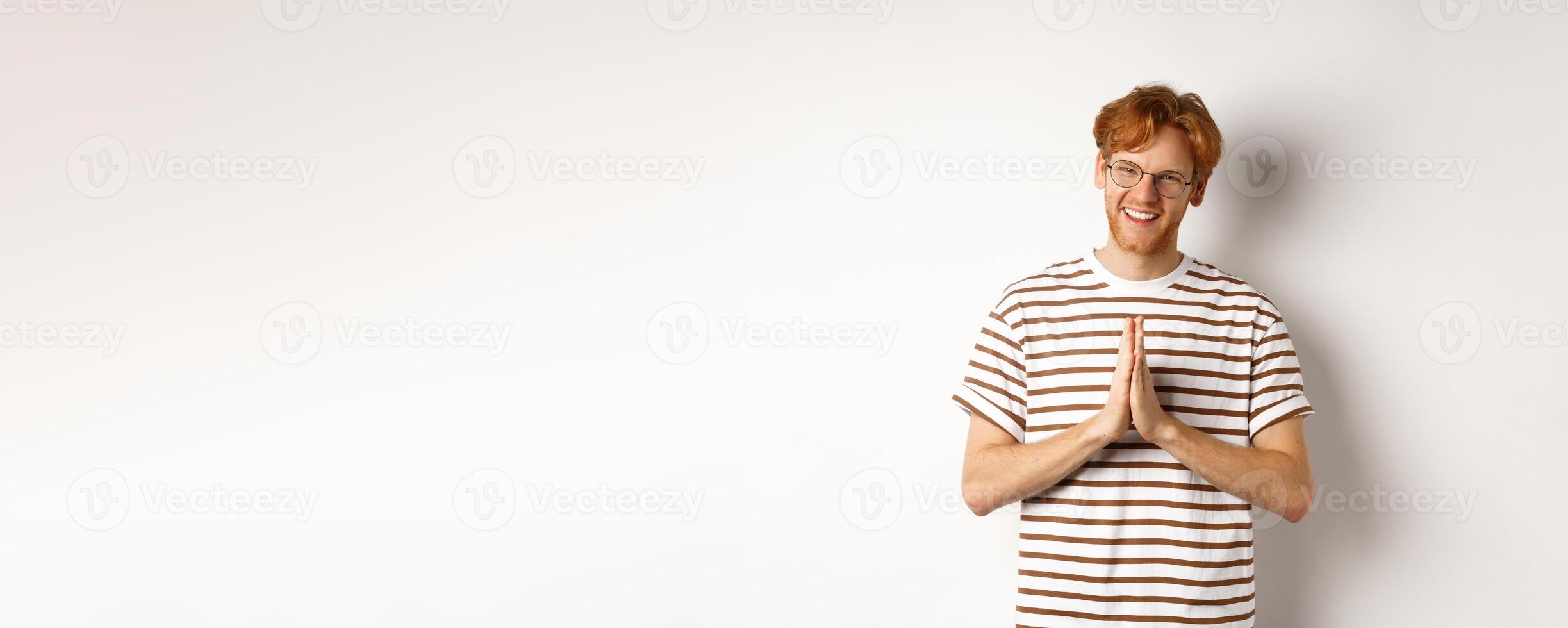 Handsome young man in glasses, red hair, showing namaste gesture and smiling, thanking you, standing grateful over white background photo