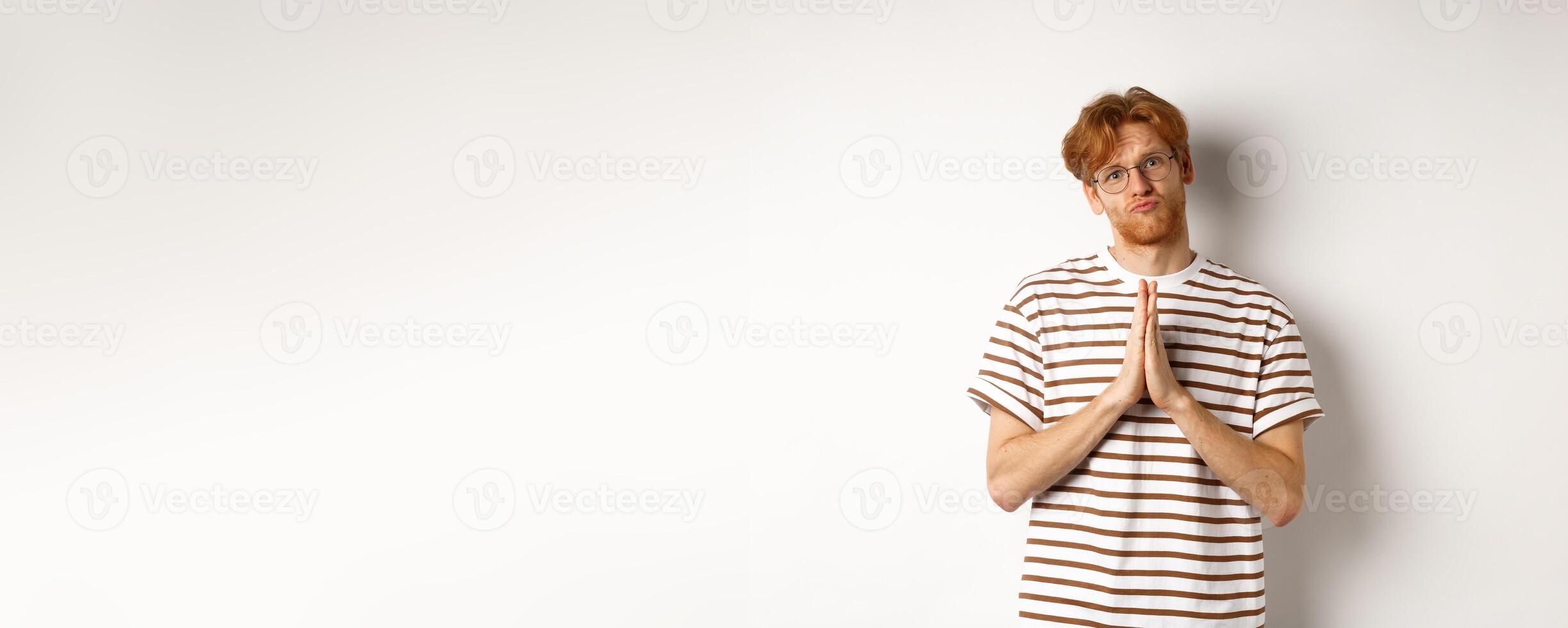 Silly redhead man asking for help, holding hands in pleading gesture and pucker lips, say please while begging you, standing over white background photo