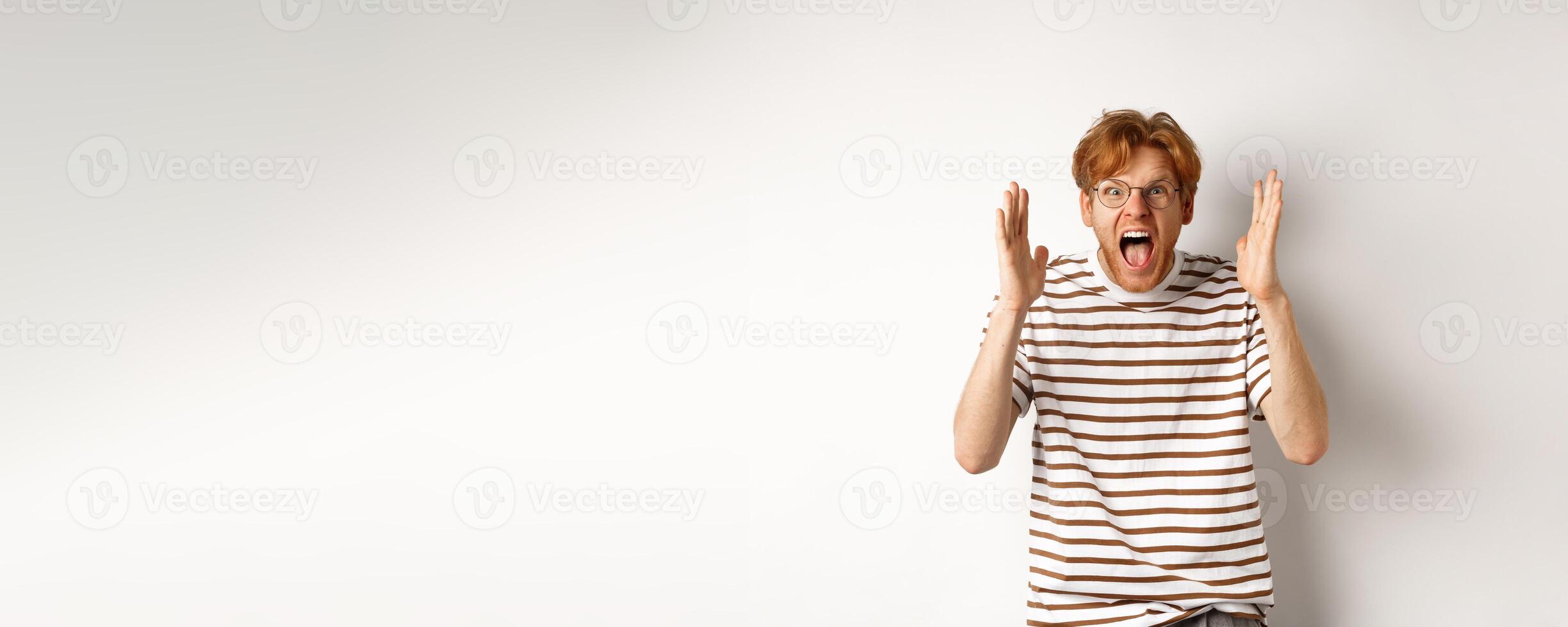 Angry young man with red hair shouting at camera, screaming and looking outraged, shaking hands, standing over white background photo