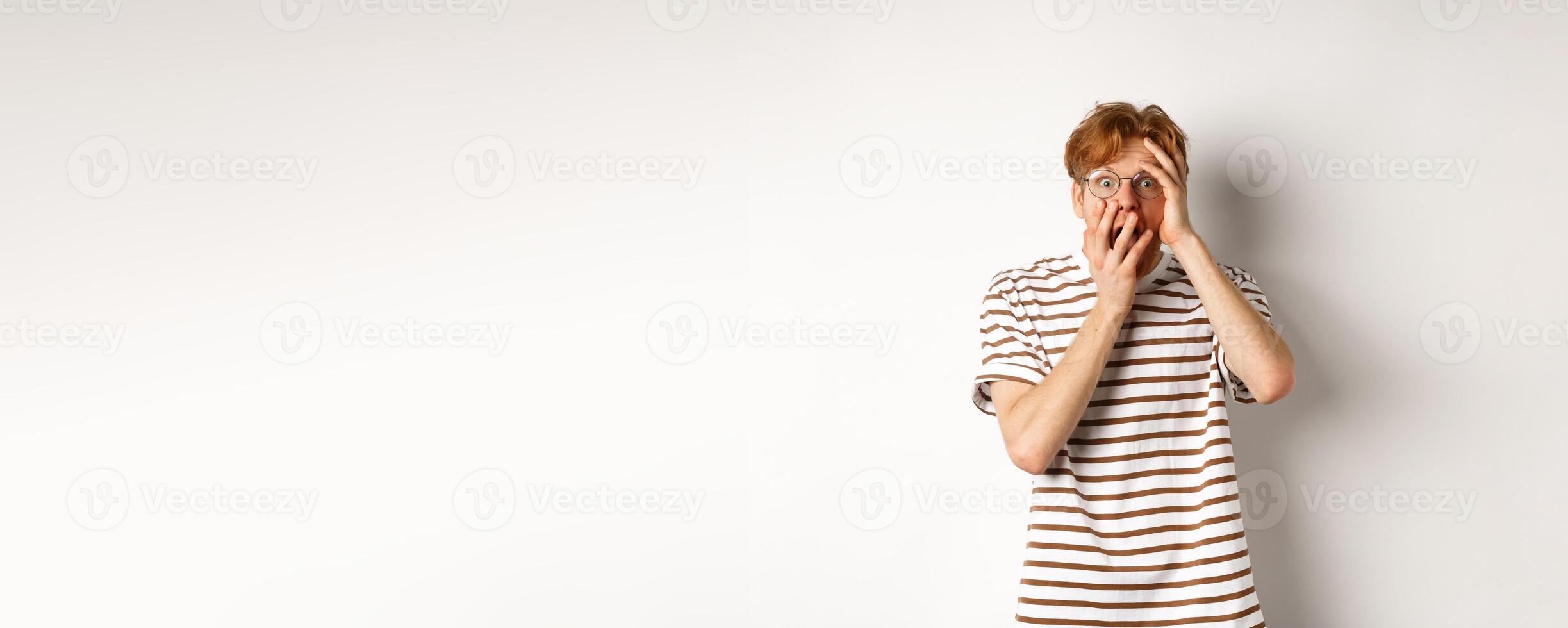 Scared redhead guy screaming terrified, holding hands on face from fear, standing over white background photo