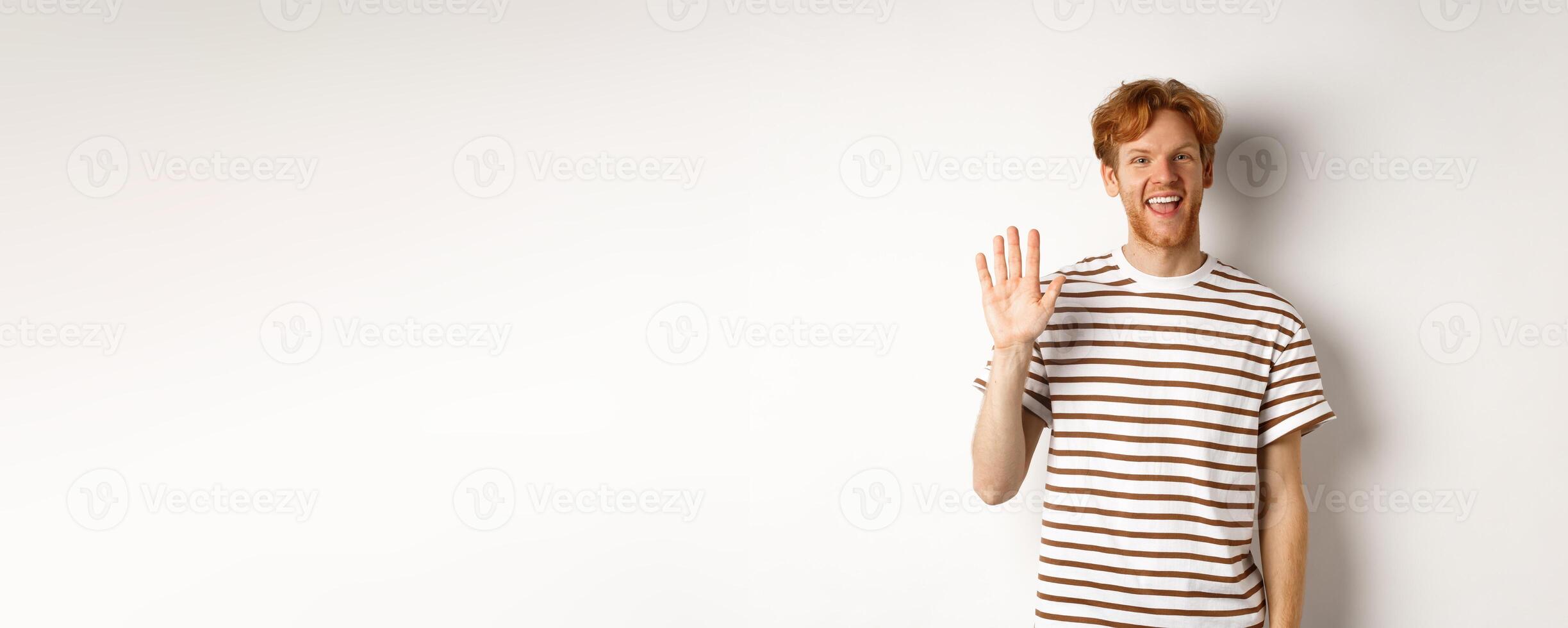 Friendly man with red hair and beard saying hi, waving hand and smiling, standing over white background photo