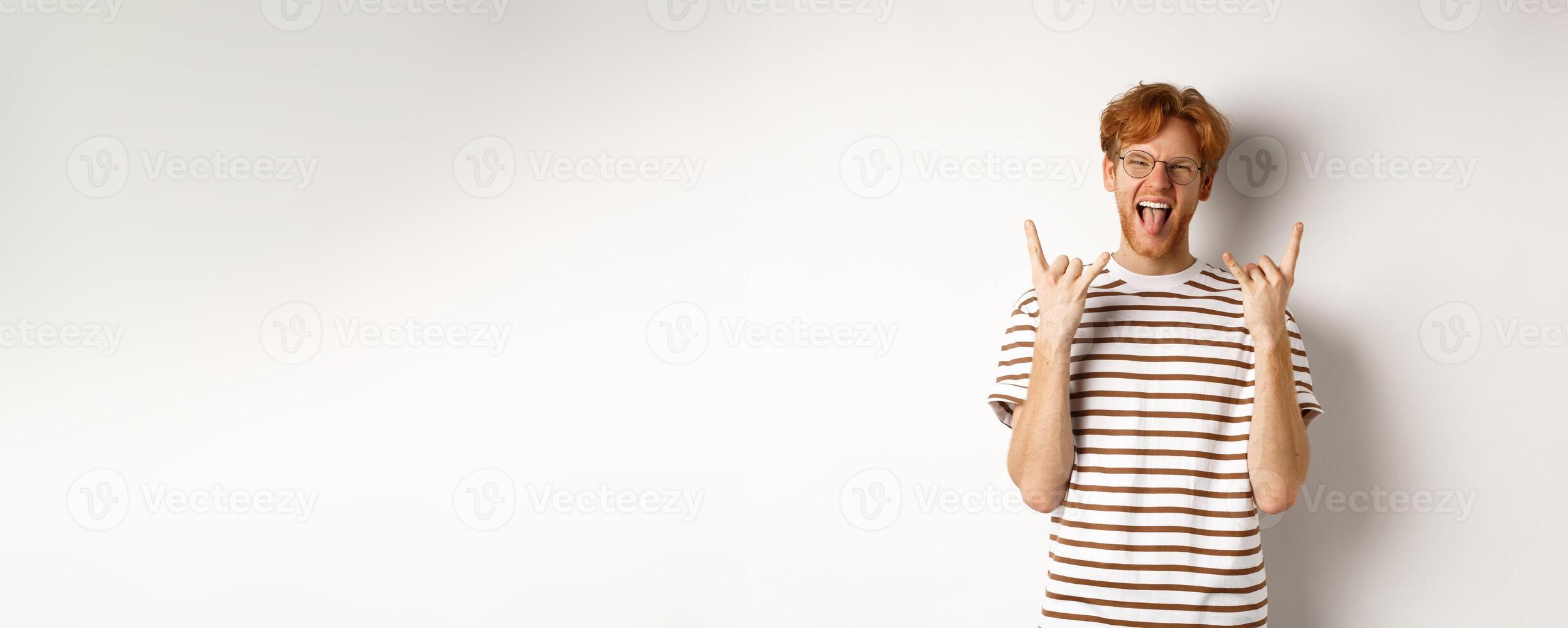 Funny and happy redhead man having fun, showing rock-n-roll horn and sticking tongue, enjoying party, standing over white background photo