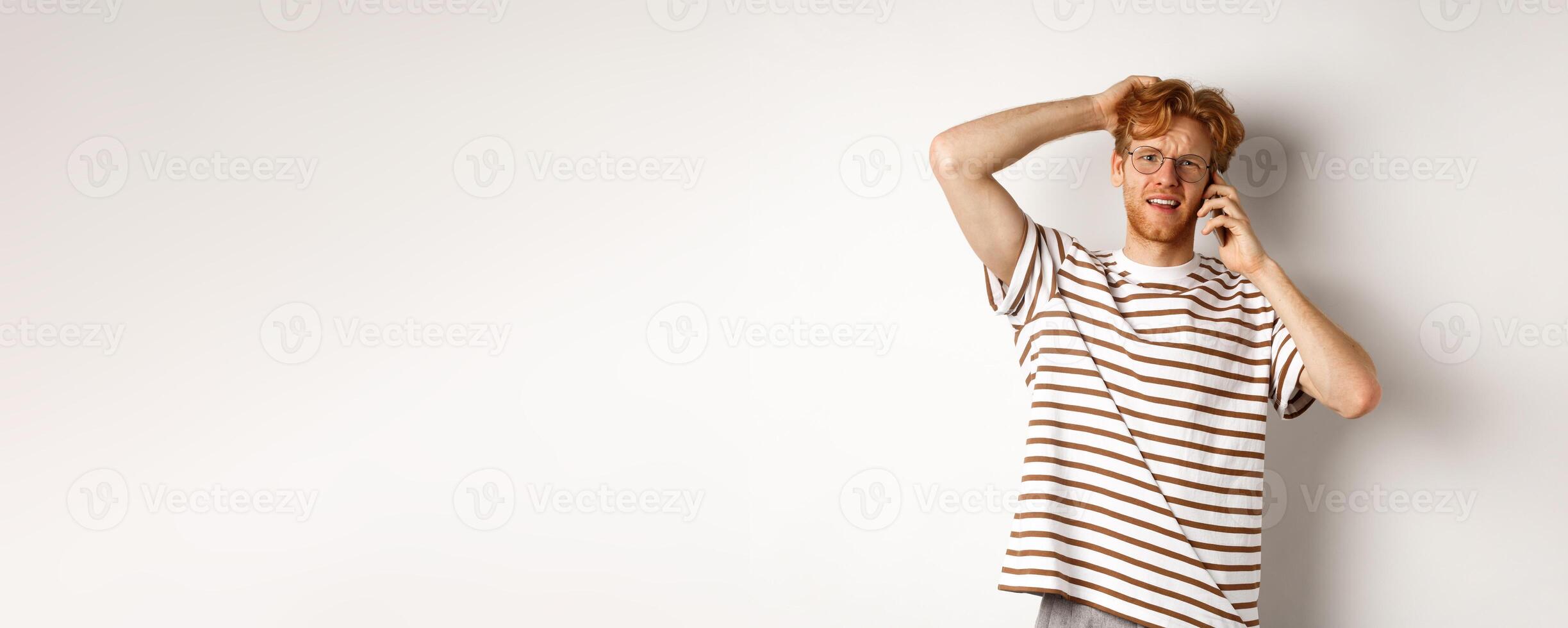 Puzzled redhead guy talking on phone, scratching head and looking confused or indecisive, standing over white background photo