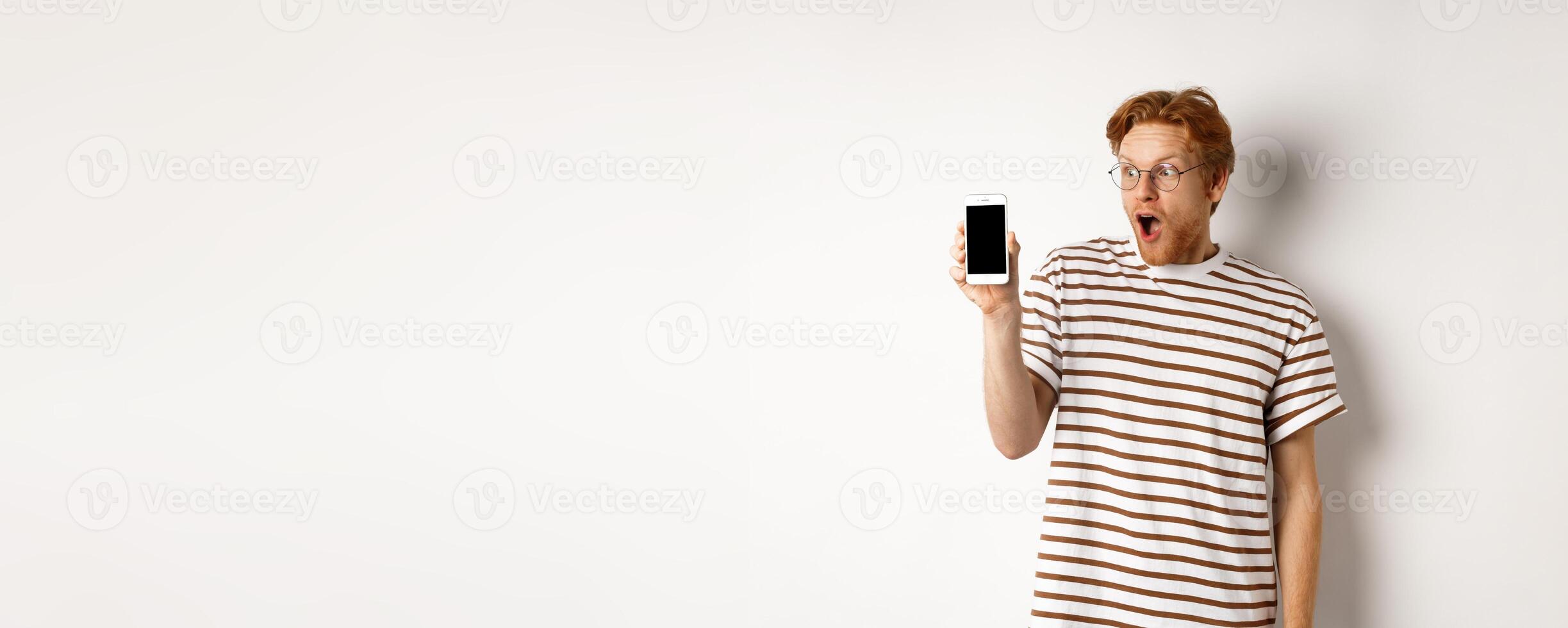 Technology and e-commerce concept. Surprised and shocked redhead guy checking out online promotion, showing blank smartphone screen and drop jaw, white background photo