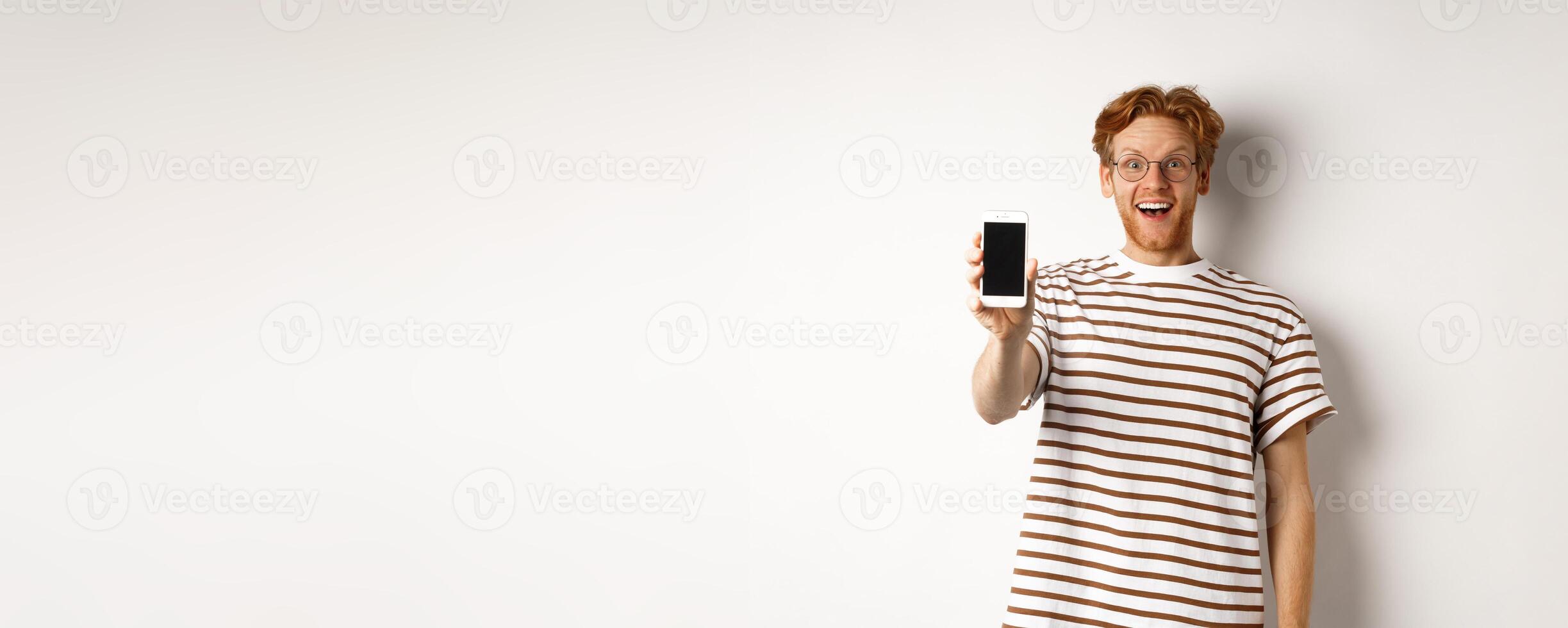 Technology and e-commerce concept. Happy young redhead man in glasses showing blank smartphone screen, looking at camera amazed, standing over white background photo