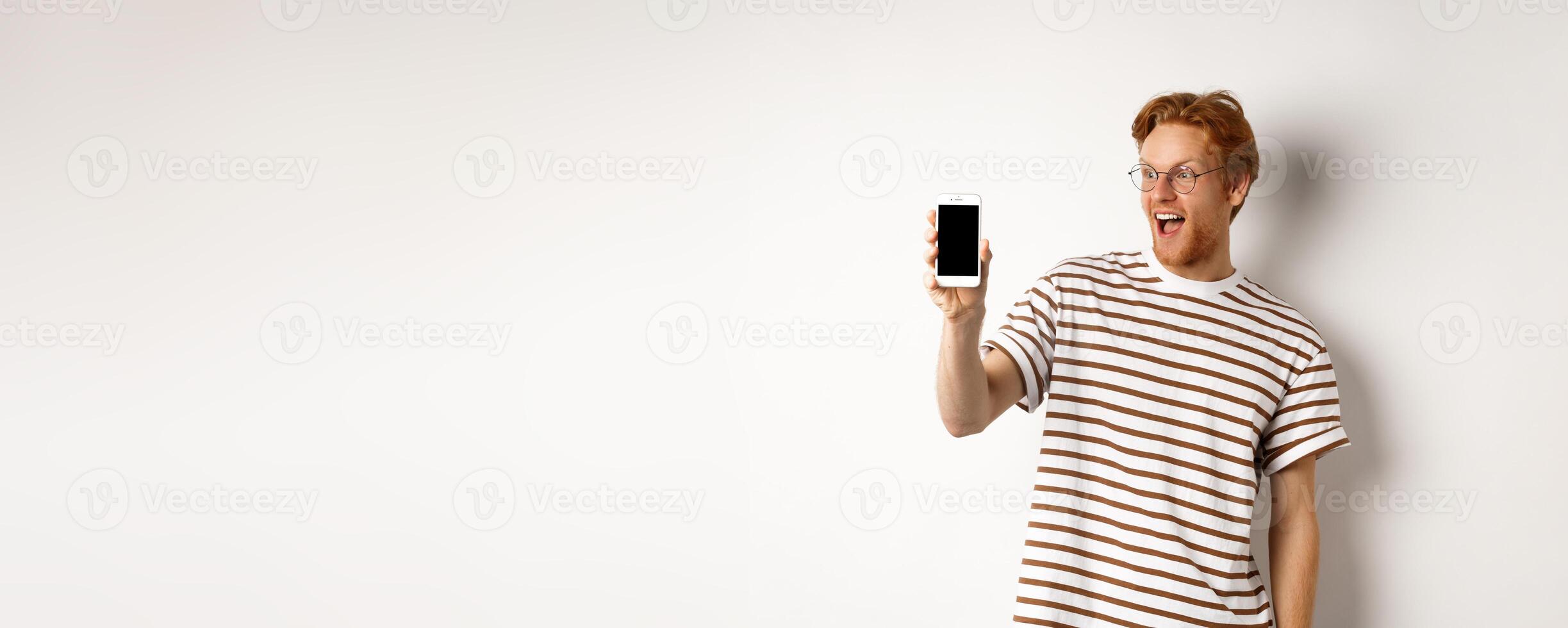 Technology and e-commerce concept. Happy young redhead man in glasses showing blank smartphone screen, looking amazed, standing over white background photo