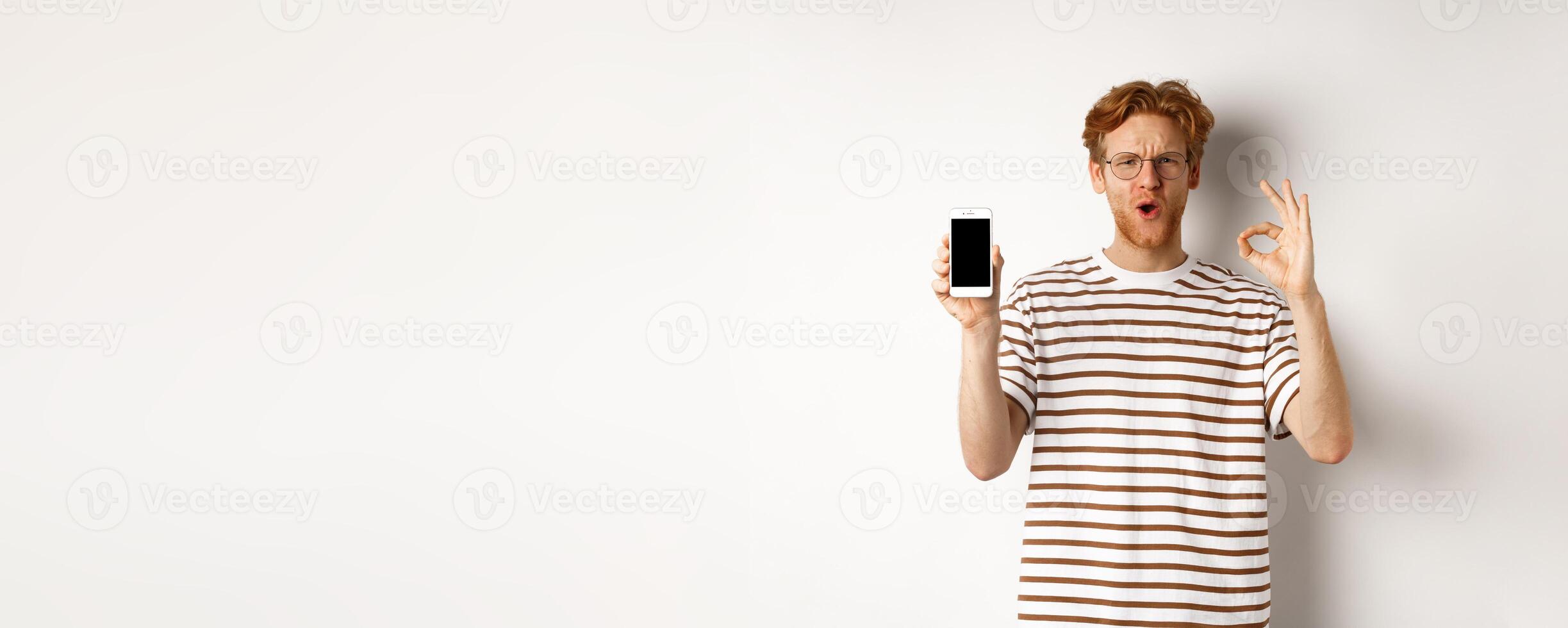 Technology and e-commerce concept. Young man with red hair showing okay sign and blank smartphone screen, praising awesome app or game, white background photo