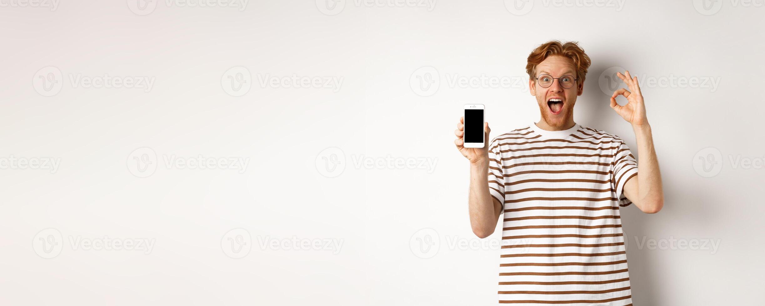 Technology and e-commerce concept. Young man with red hair showing okay sign and blank smartphone screen, praising awesome app, standing over white background photo