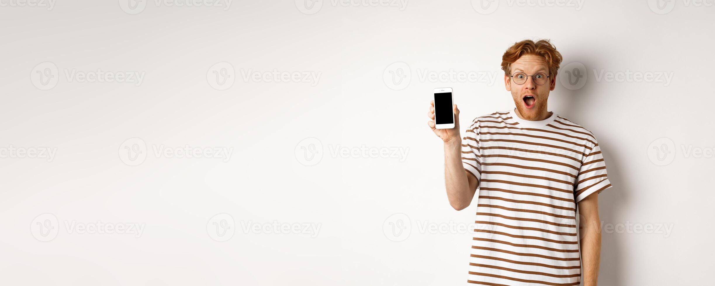 Technology and e-commerce concept. Surprised and shocked redhead guy checking out online promotion, showing blank smartphone screen and drop jaw, white background photo