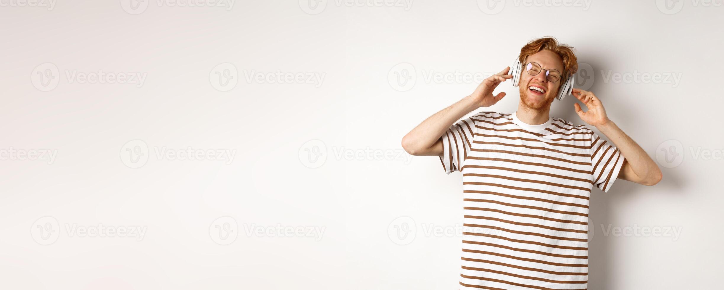 Technology concept. Happy redhead man listening music in headphones and singing along, standing over white background photo