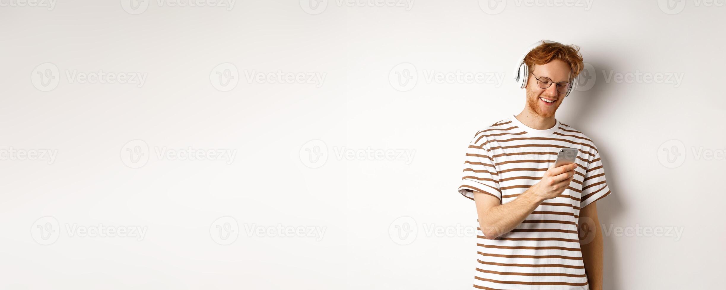 Technology concept. Young redhead hipster in glasses, listening music in headphoes, reading message on smartphone, white background photo