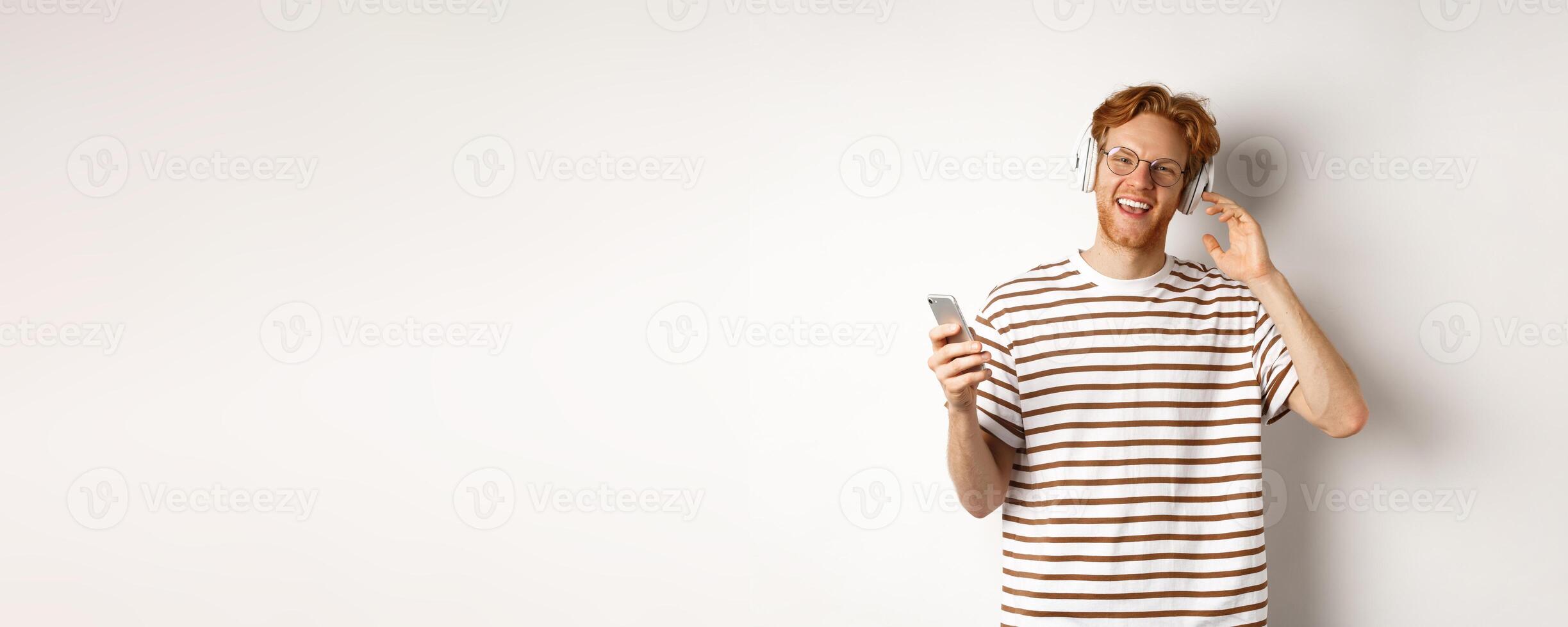 Technology concept. Young man with red hair and beard listening music in headphones and using smartphone, smiling at camera, white background photo
