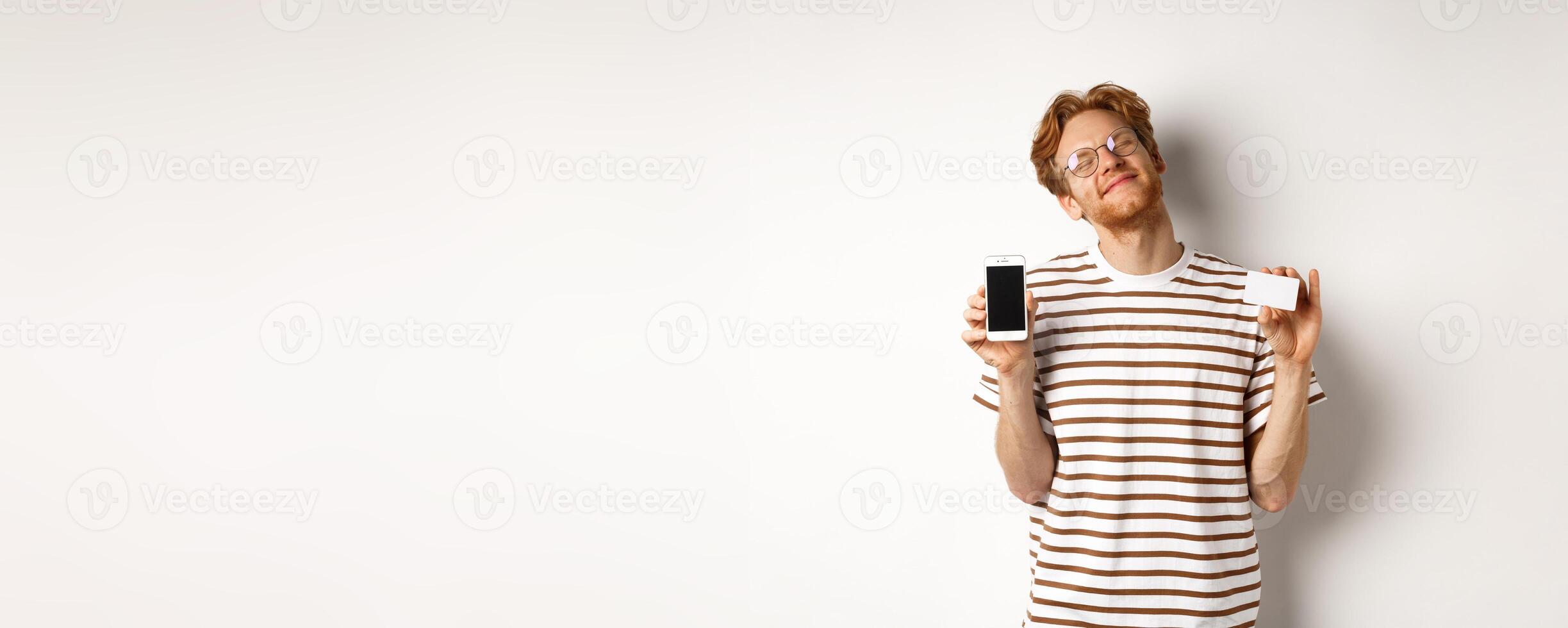 Shopping and finance concept. Pleased young man with red hair smiling from satisfaction, showing smartphone blank screen and credit card, white background photo