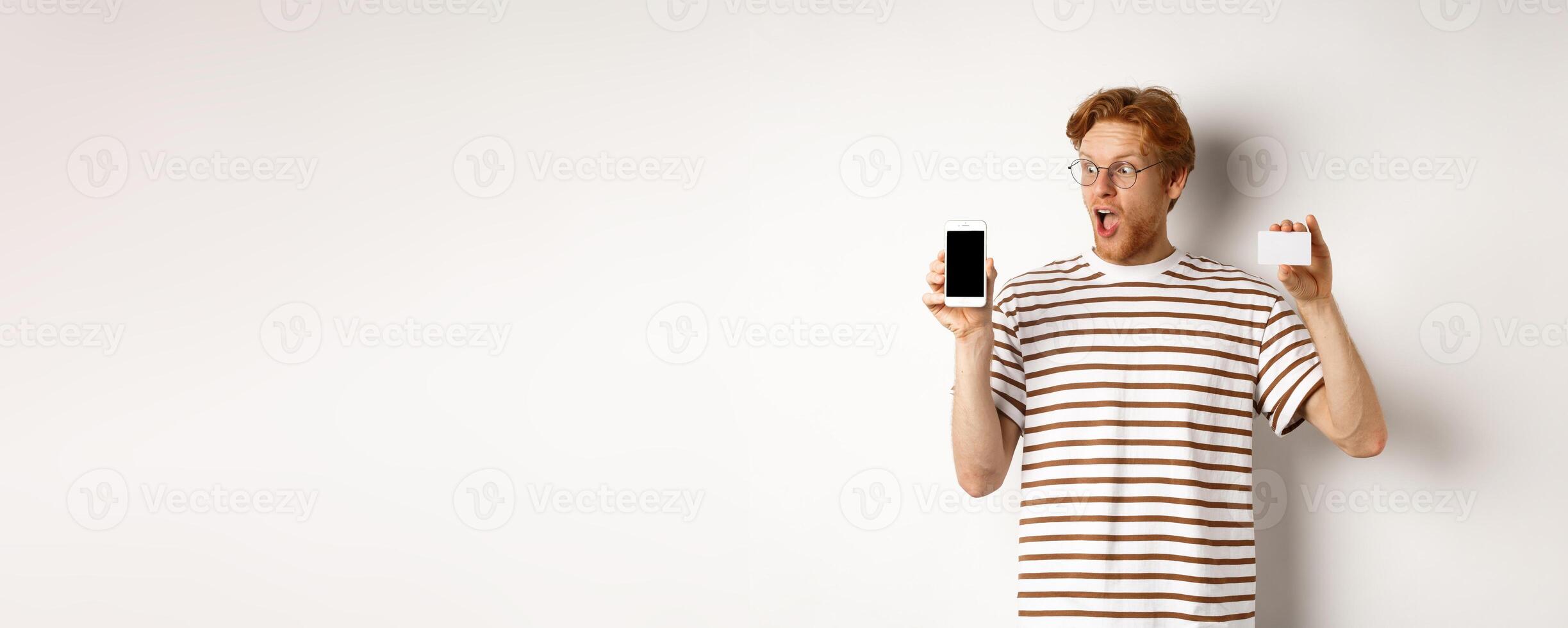 Shopping and finance concept. Amazed young man with red hair showing plastic credit card and smartphone blank screen, staring at display impressed, white background photo