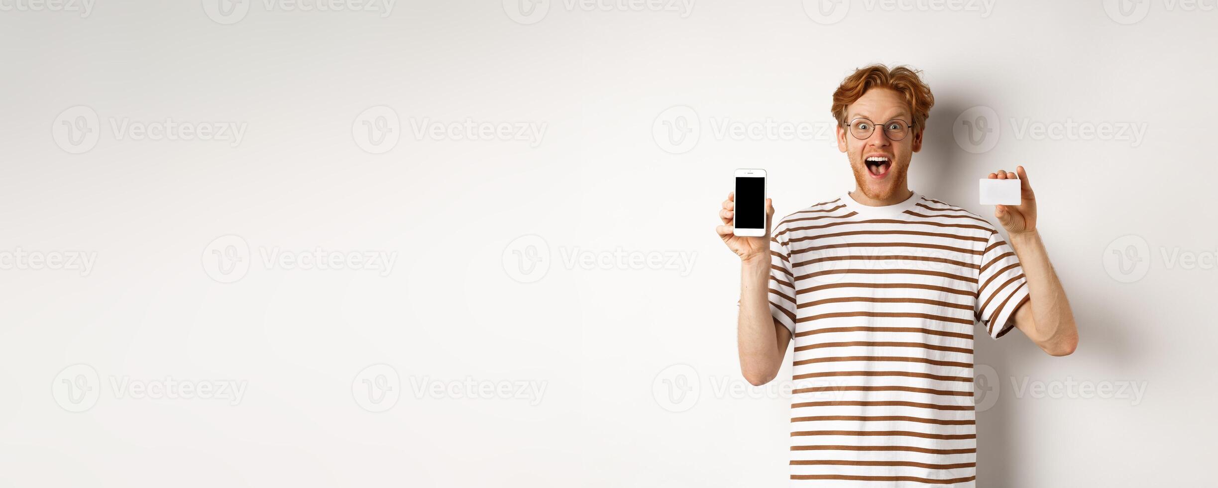 Shopping and finance concept. Young man showing blank mobile screen and plastic credit card, smiling at camera, white background photo