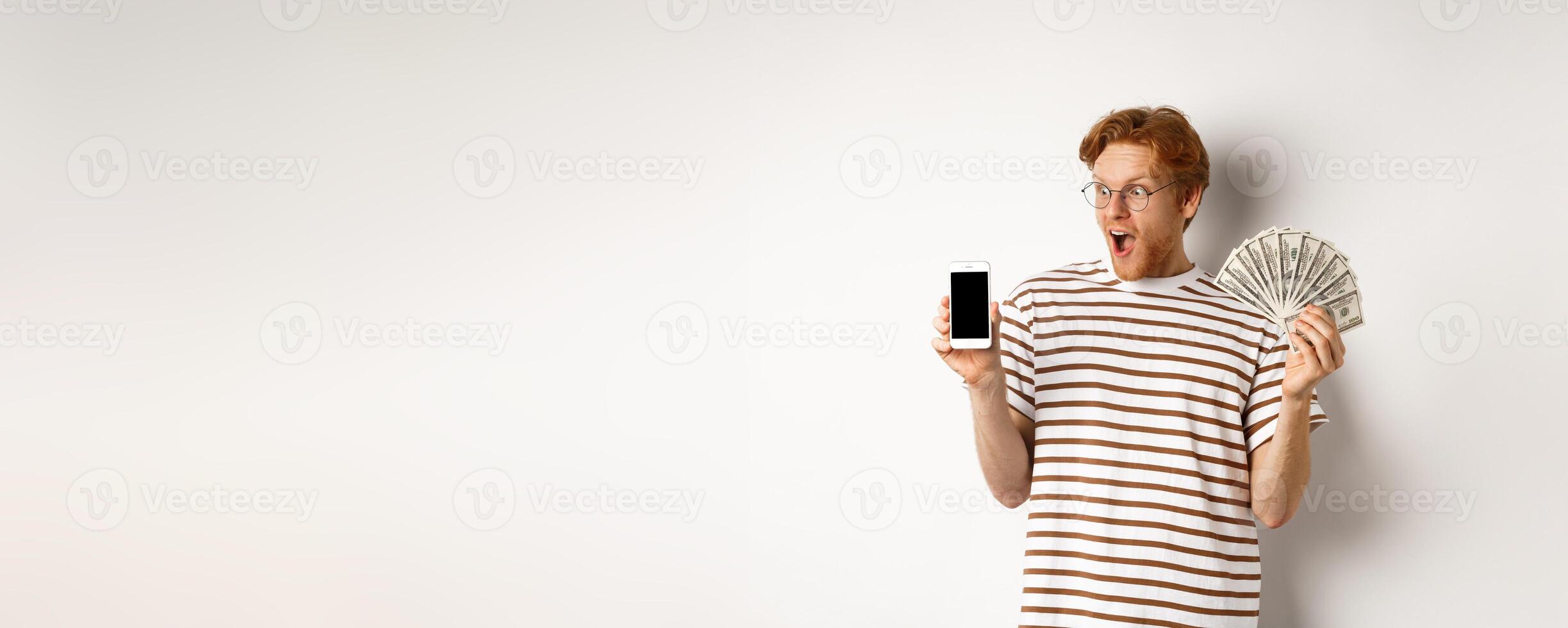 Amazed redhead man showing smartphone app on blank screen and money, winning prize cash online, standing over white background photo