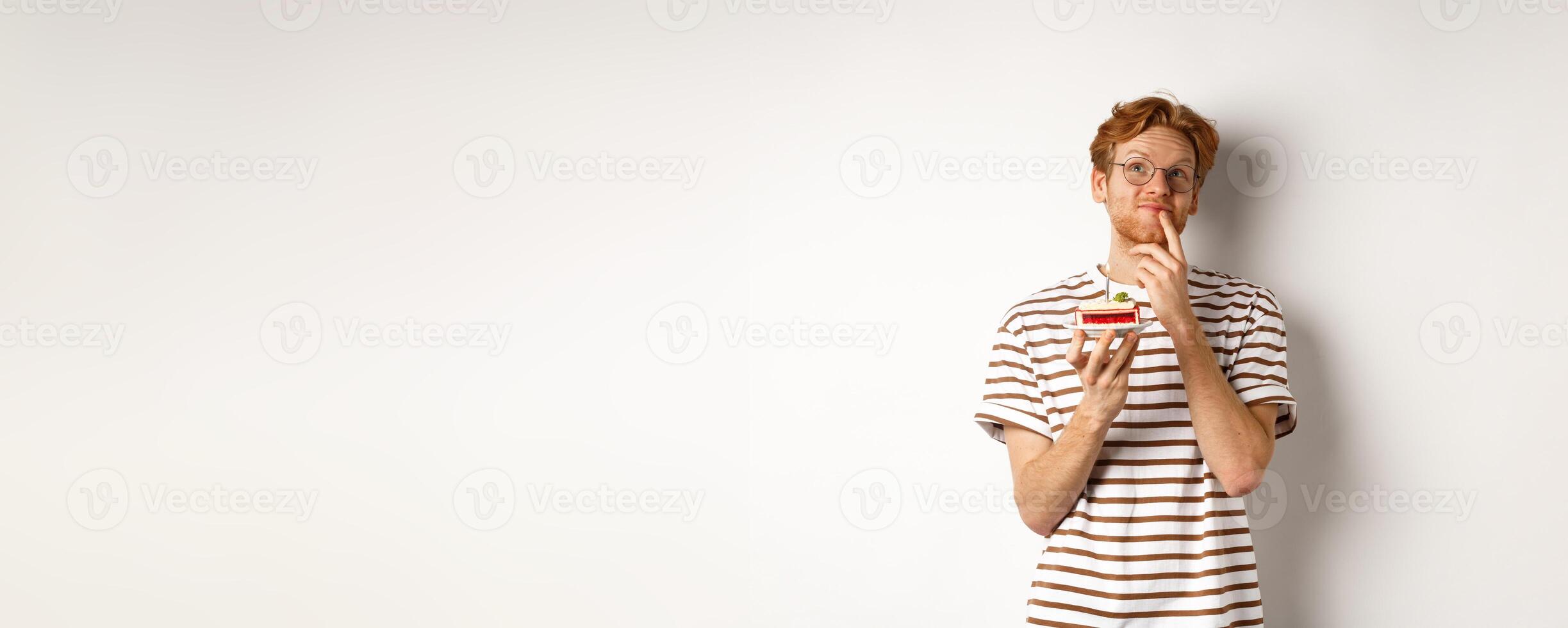 Holidays and celebration concept. Happy young man with red hair and glasses having birthday, holding cake with candle and thinking of b-day wish, standing over white background photo
