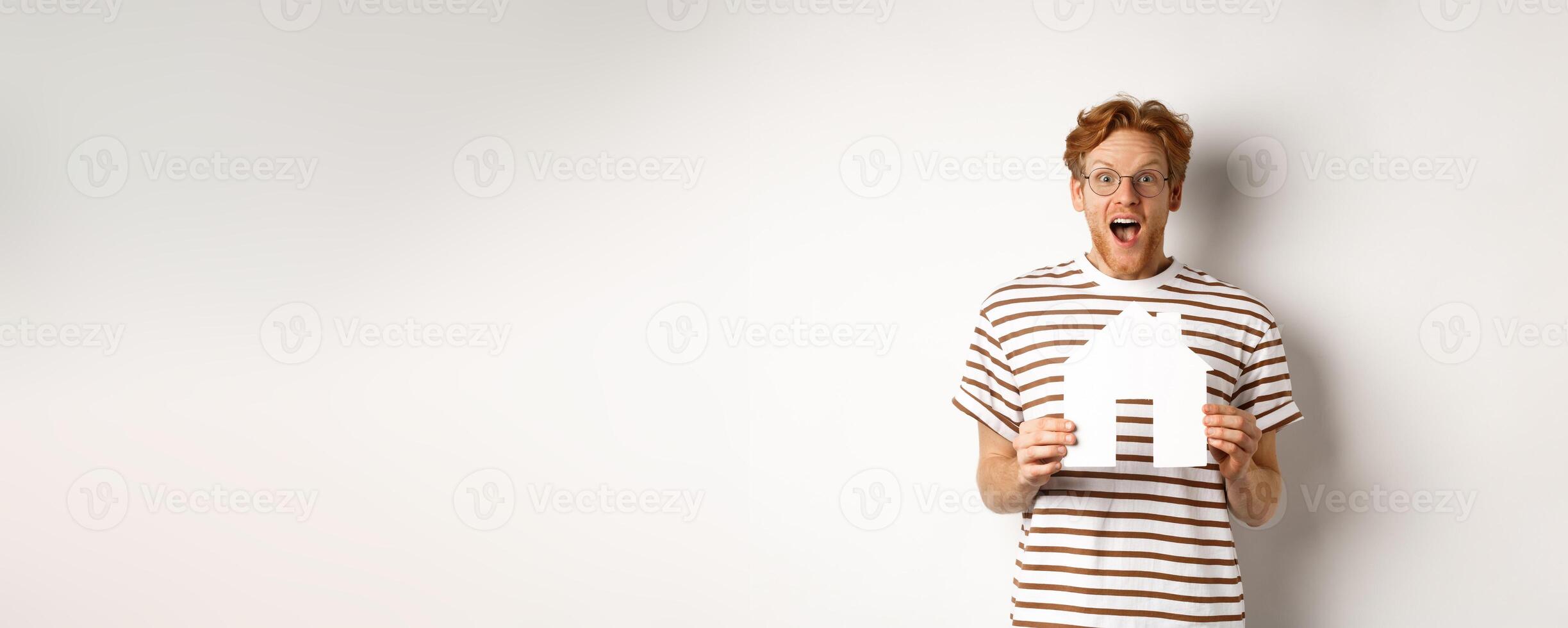Happy and surprised redhead man winning house, holding paper home model and staring at camera, standing joyful over white background photo