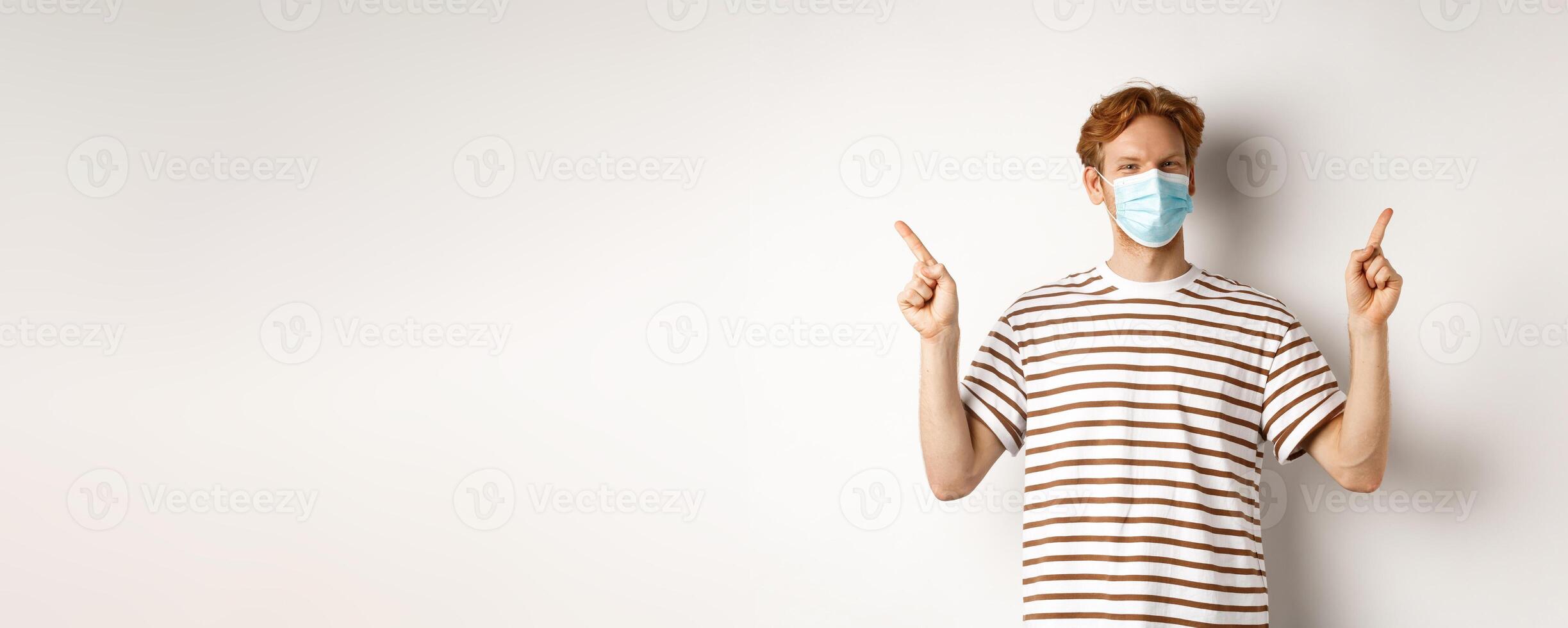 Covid, virus and social distancing concept. Handsome young man with red hair, wear face mask and pointing sideways at two promo offers, white background photo