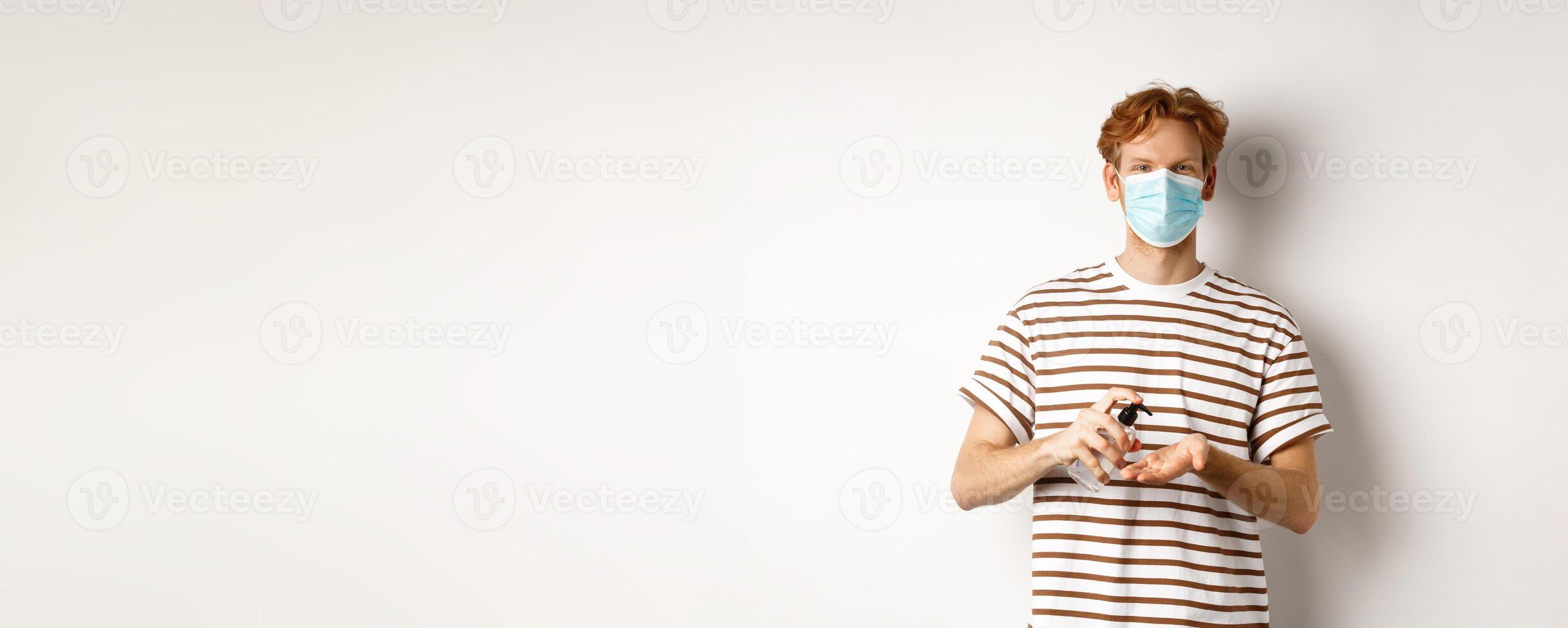 Covid, virus and social distancing concept. Teenager redhead guy in face mask clean hands with sanitizer, using antiseptic, standing over white background photo