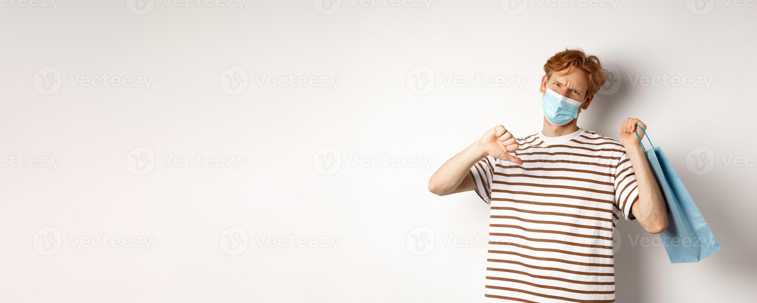 Concept of social distancing and shopping. Disappointed young man with red hair, wearing face mask, holding shopping bag and showing thumb-down from dislike, disapprove store photo