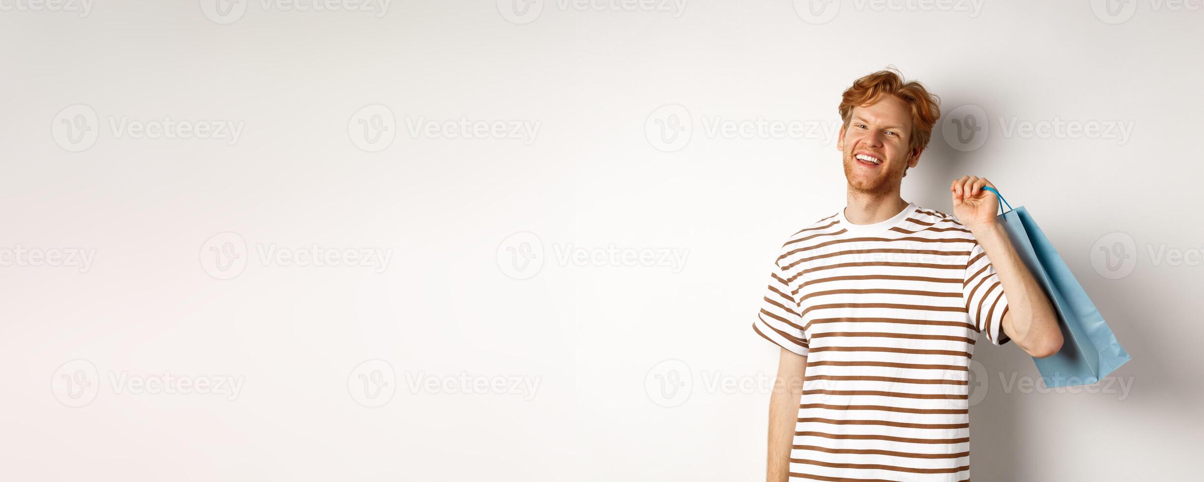 Cheerful bearded man with red hair enjoying shopping, holding bag over shoulder and smiling, standing over white background photo