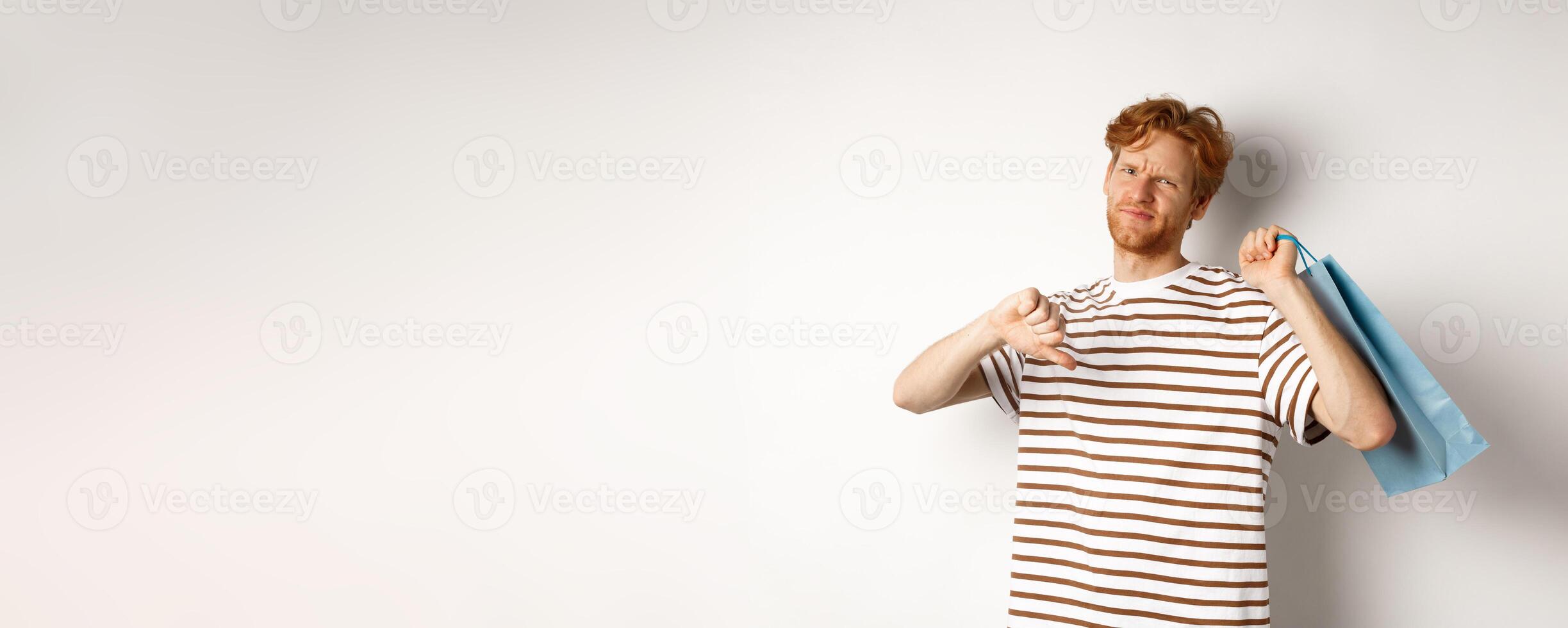 Disappointed young man with red hair and beard showing thumbs-down after bad shopping experience, holding bag over shoulder and frowning upset, white background photo