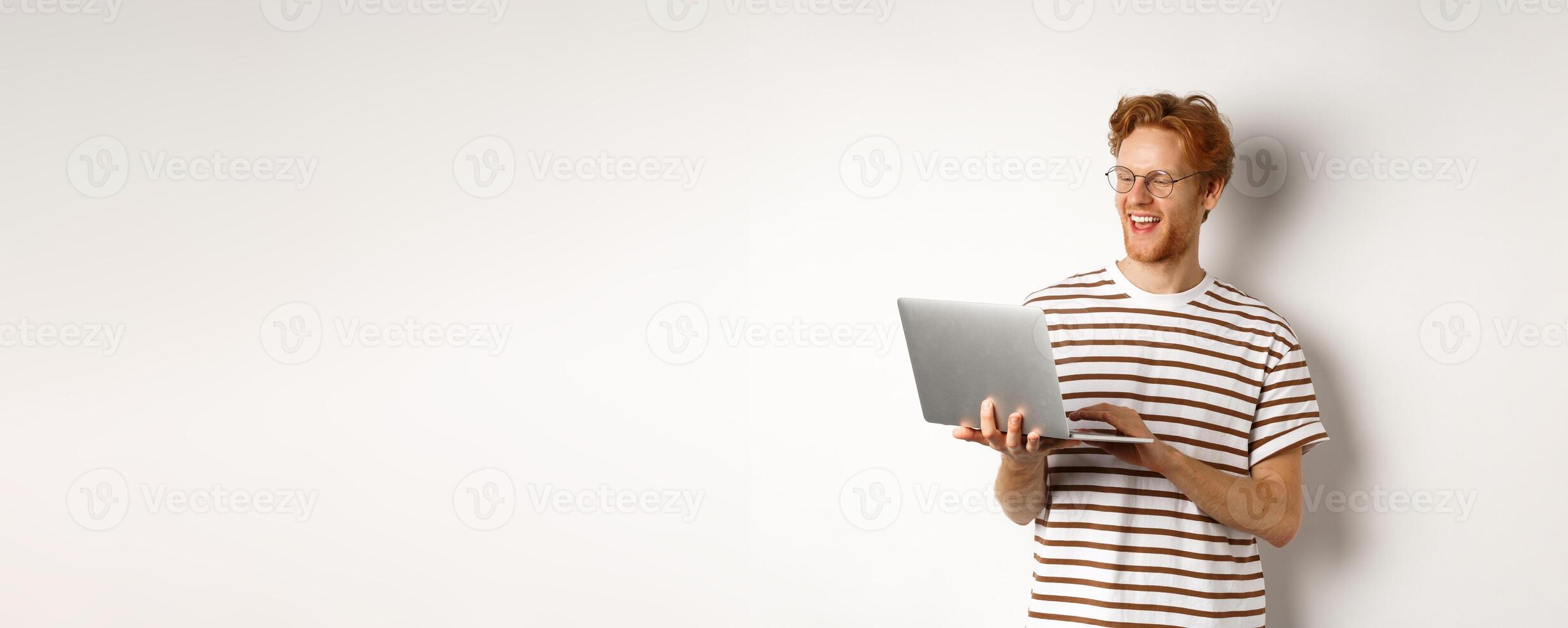 Handsome redhead man working freelance on laptop, smiling and looking pleased at computer screen, white background photo