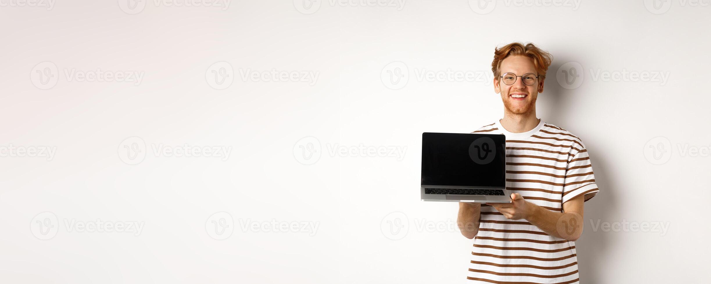Smiling proud redhead guy showing empty laptop screen. Handsome young man demonstrating logo on computer display, white background photo