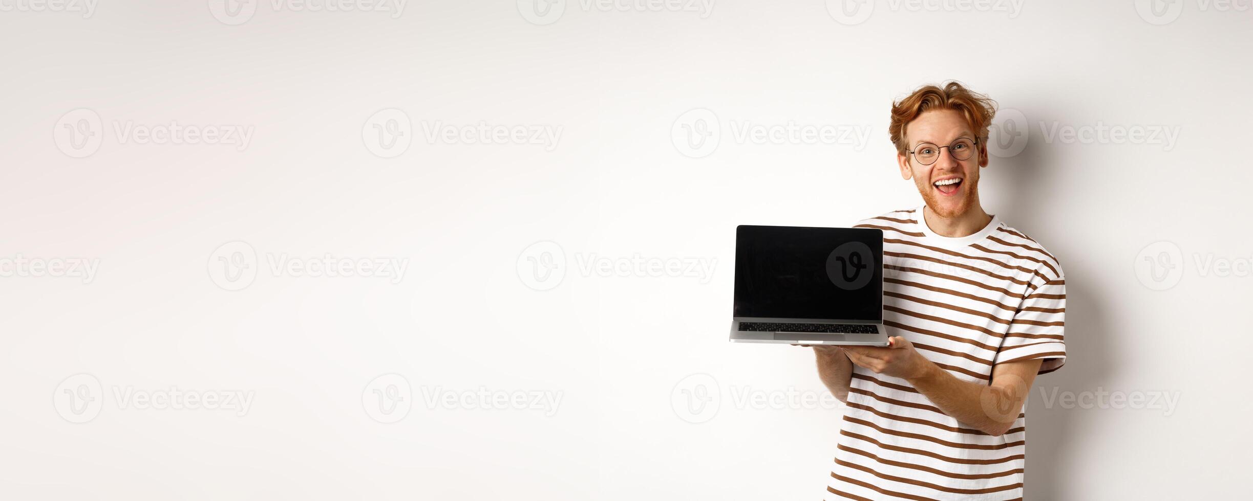 Funny redhead man in glasses showing laptop screen advertisement and smiling. Guy with red hair demonstrates promo or banner on display, white background photo
