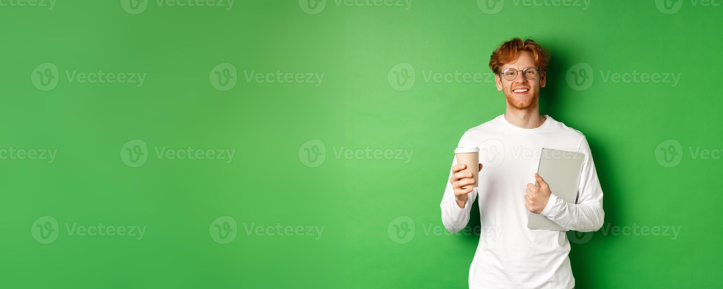 Handsome young man, employee with laptop drinking coffee on break, smiling satisfied, standing over green background photo