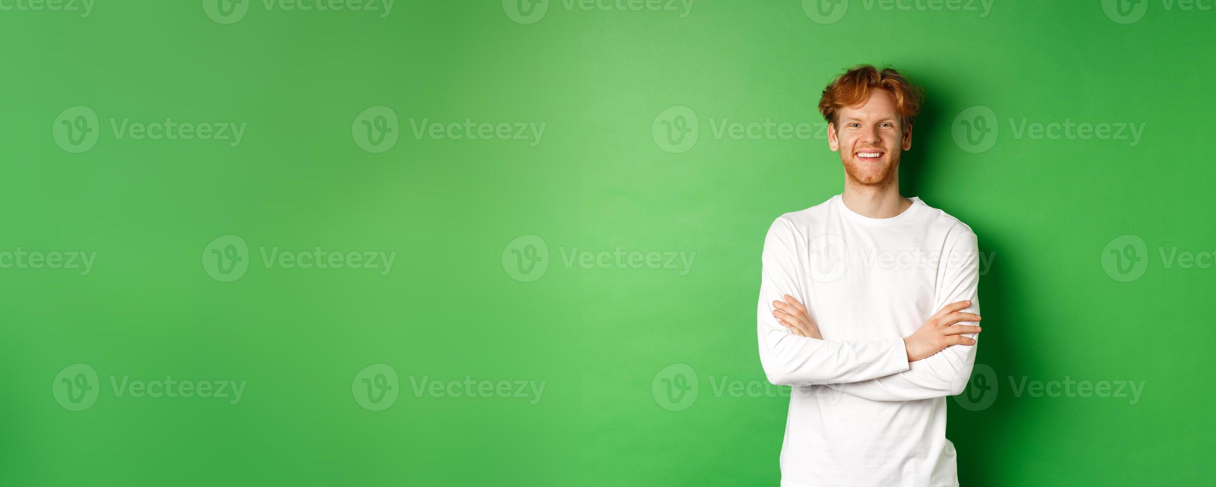 Young ambitious man with red hair standing over green background, holding hands crossed on chest and smiling photo