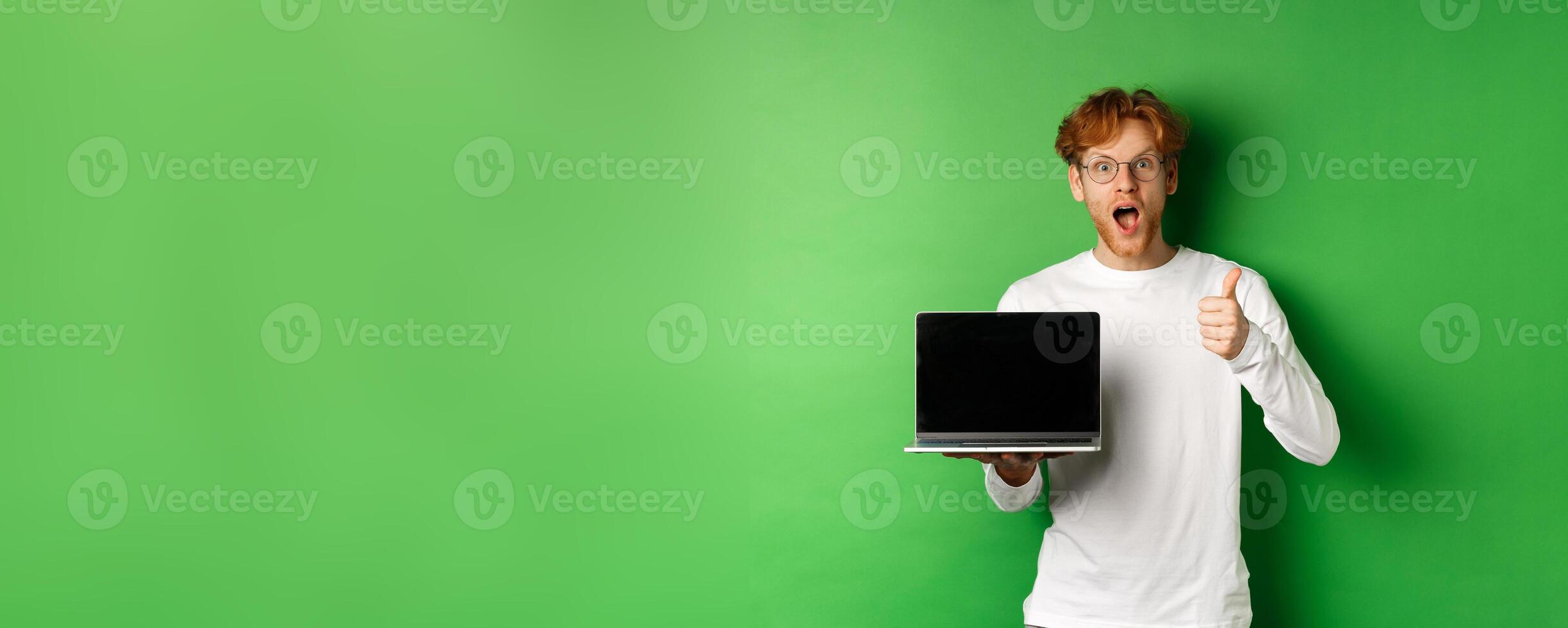Impressed caucasian man with red hair and beard, showing blank laptop screen and thumb-up, looking amazed at camera, praising something online, green background photo