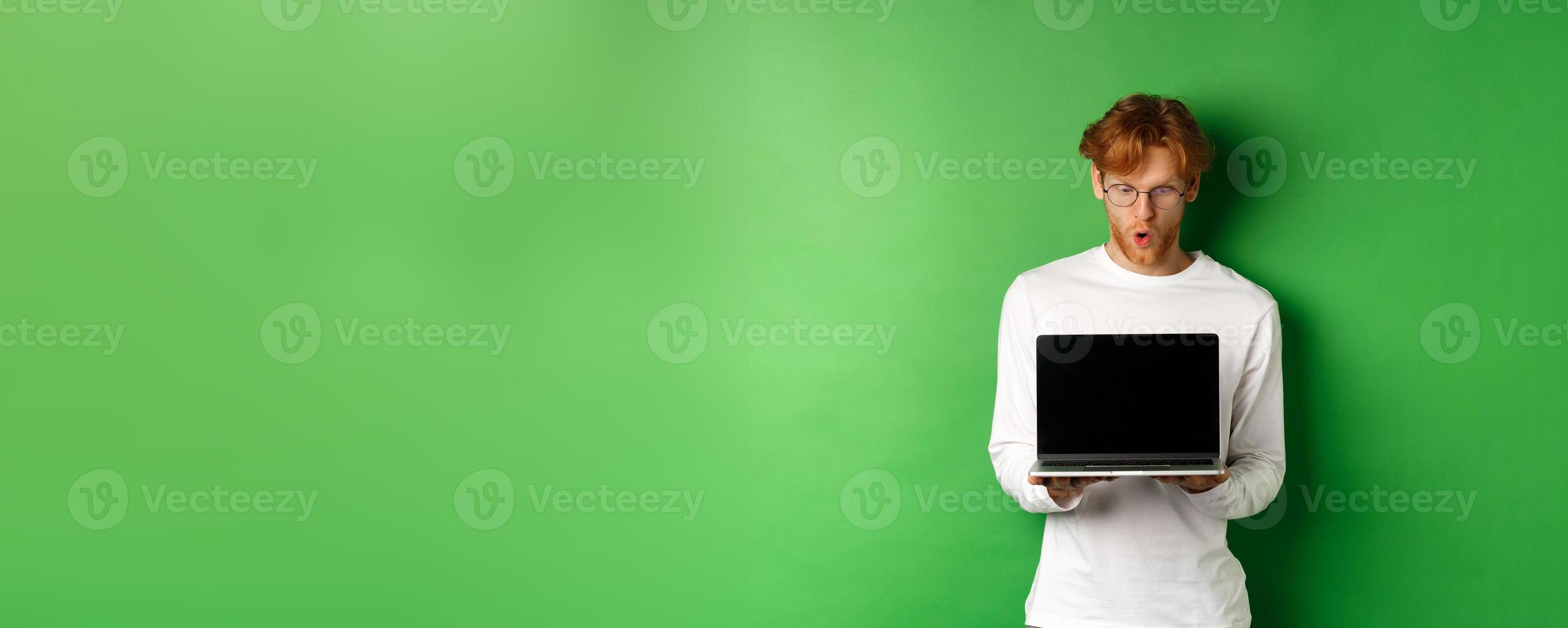 Impressed redhead nerdy guy in glasses showing blank laptop screen and looking at display amazed, standing over green background photo