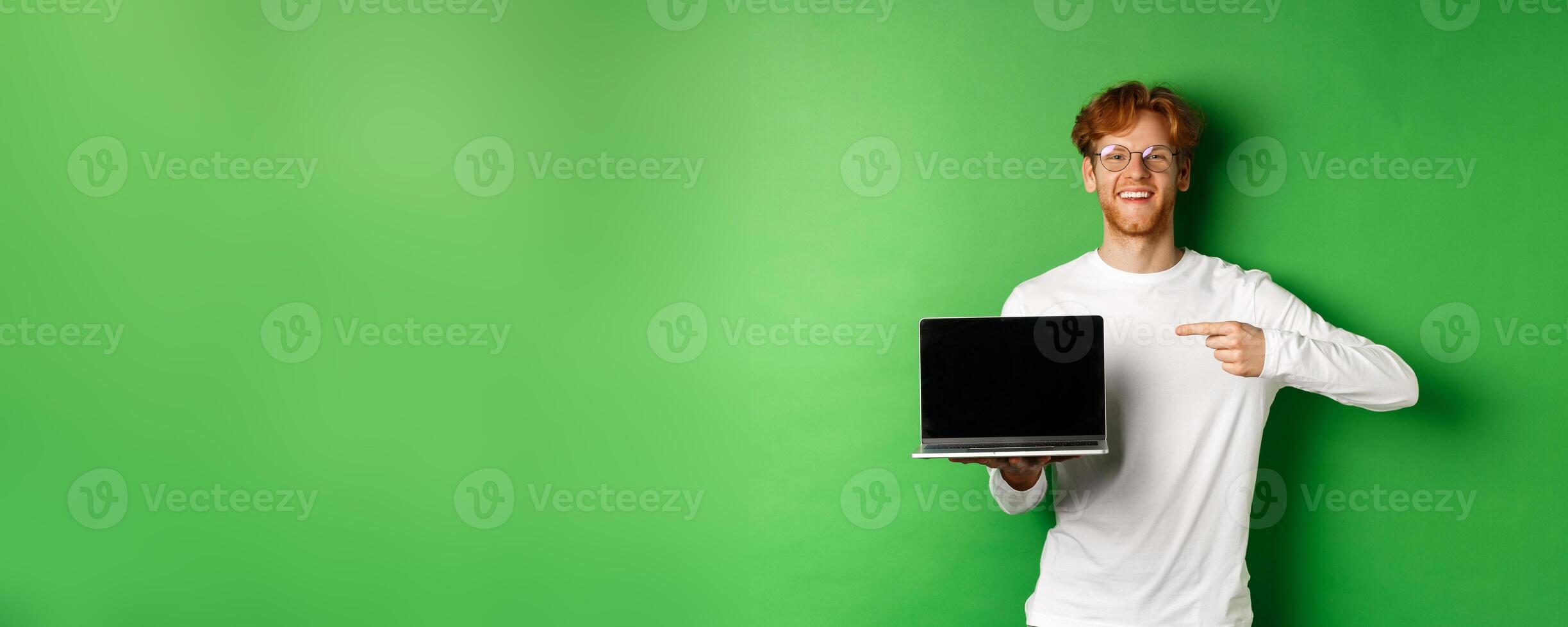 Happy redhead guy in glasses and white long-sleeve t-shirt, pointing finger at blank laptop screen and smiling, standing over green background photo