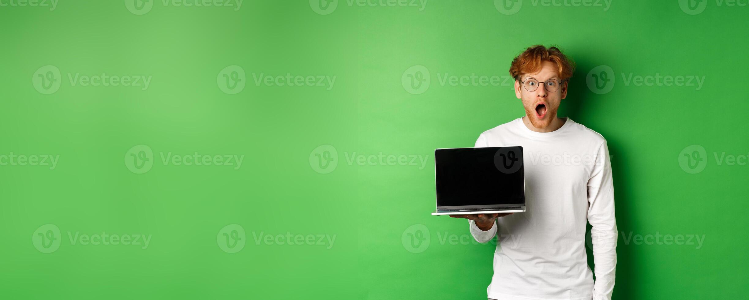 Impressed redhead guy in glasses drop jaw after seeing online promo, showing laptop screen and staring at camera with disbelief, standing over green background photo
