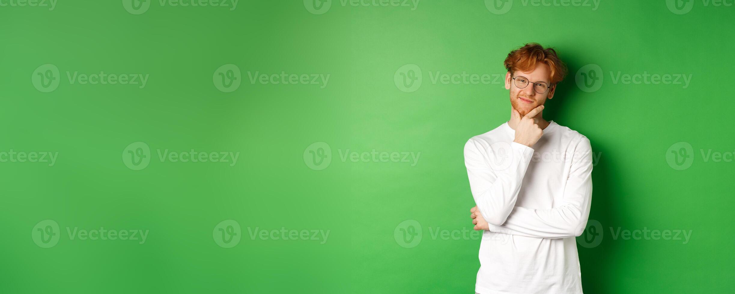 Intrigued young man with red hair, wearing glasses, looking at camera pleased and thoughtful, having an idea, standing over green background photo