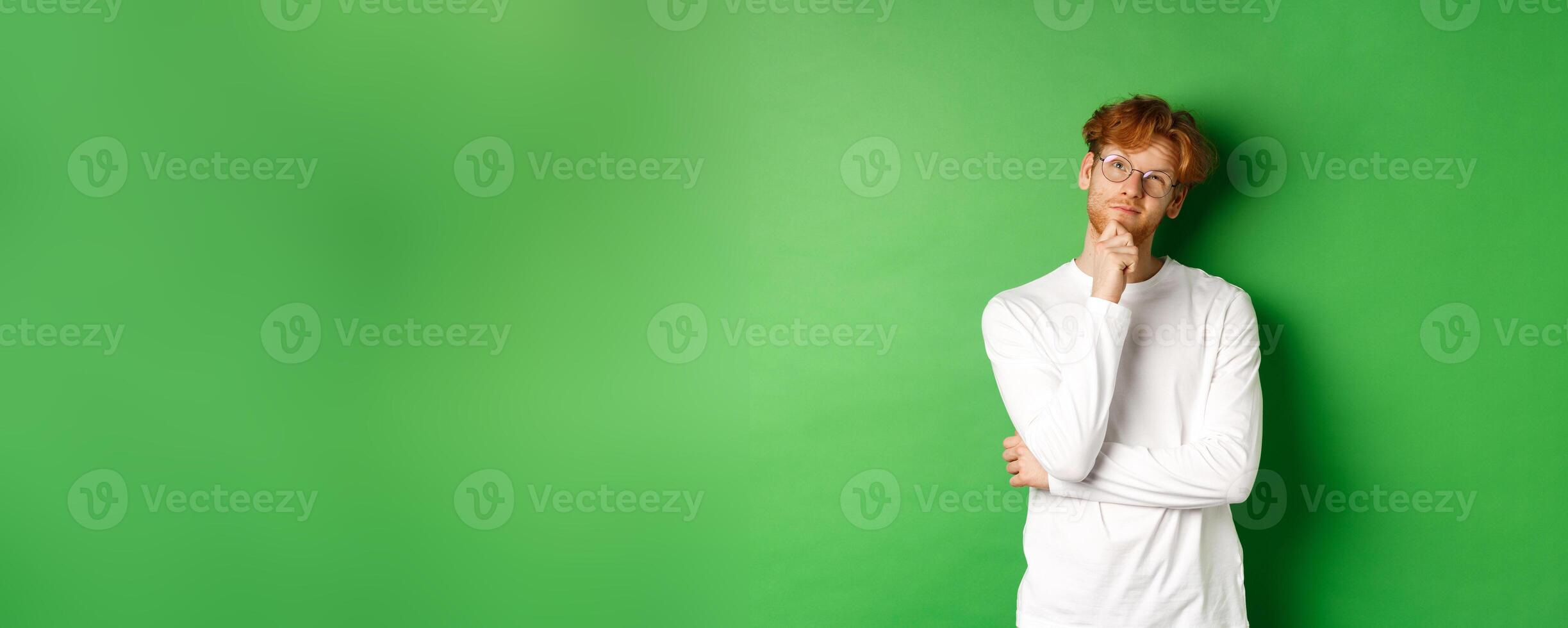 Thoughtful redhead man in glasses making choice, looking up and thinking, standing over green background photo
