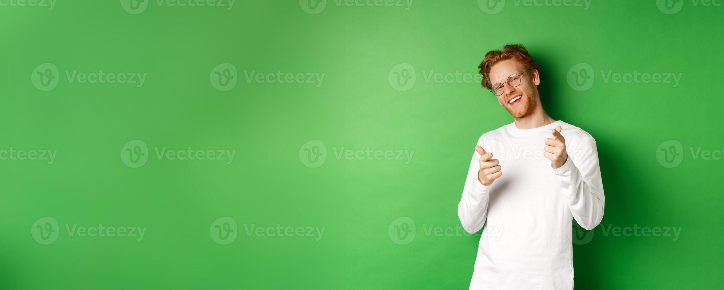 Cheeky young man in glasses praising and congratulating you, pointing fingers at camera and smiling, standing in glasses and white long-sleeve against green background photo