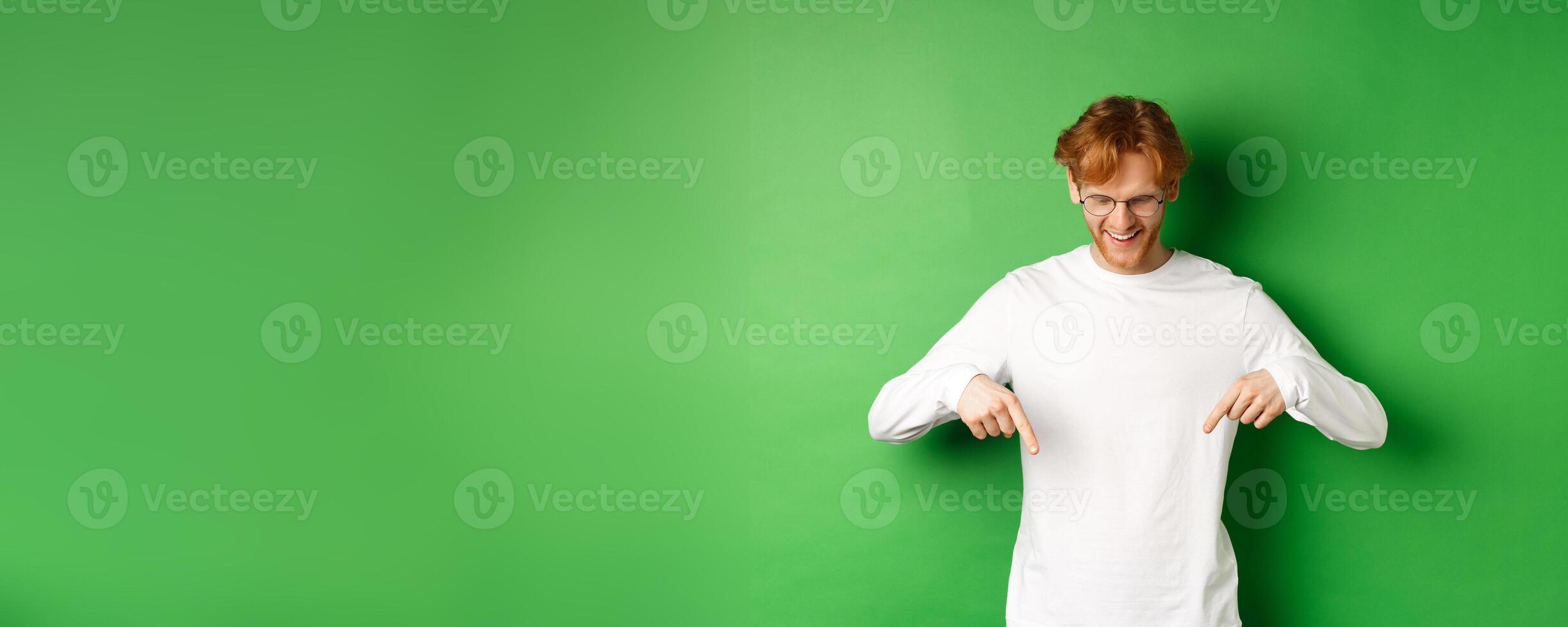 Handsome redhead man in glasses and white long-sleeve checking out advertisement, pointing fingers and looking down with pleased smile, standing over green background photo