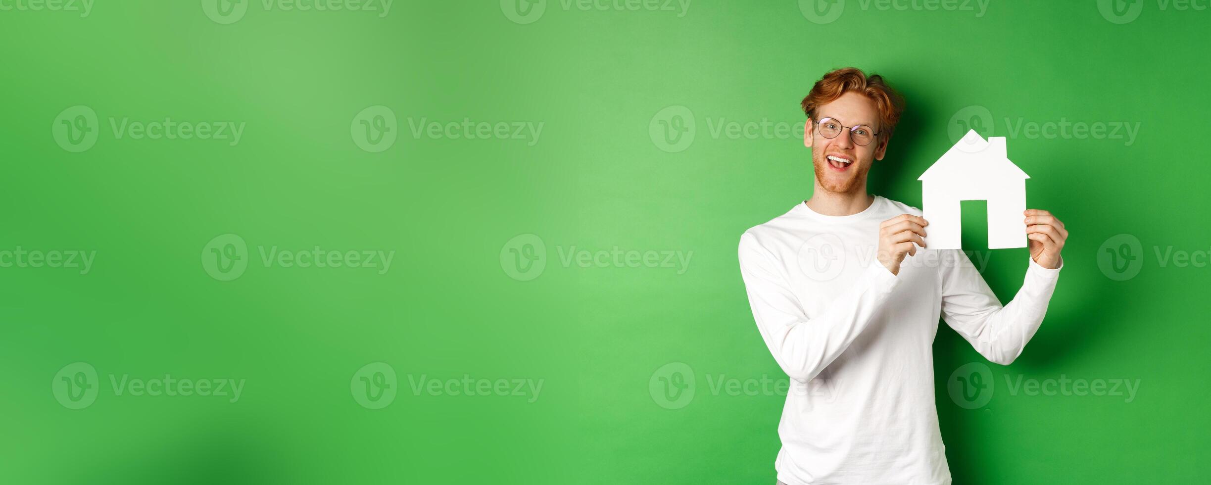Real estate and buying property concept. Handsome young man with red hair showing house cutout, searching for new home, standing over green background photo