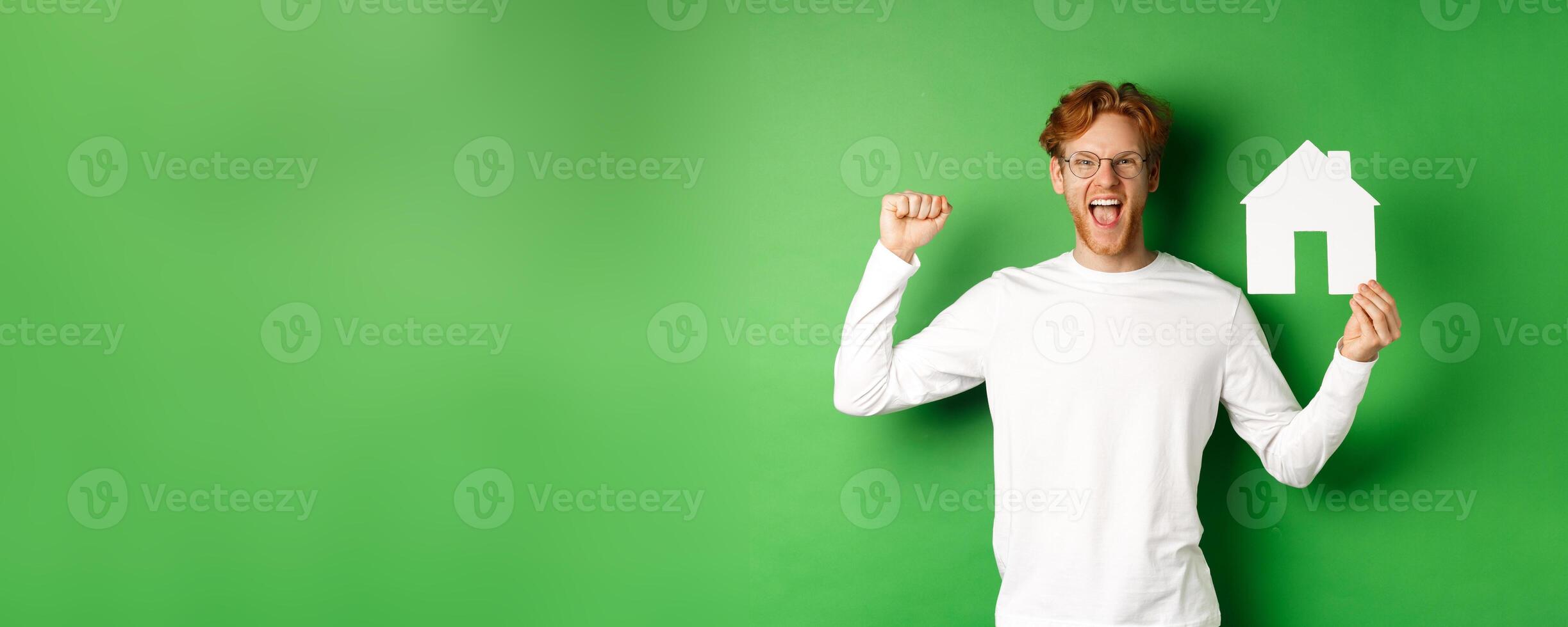 Real estate. Happy caucasian man in glasses with red hair celebrating, showing paper house cutout and making fist pump sign, shouting for joy, buying property, green background photo