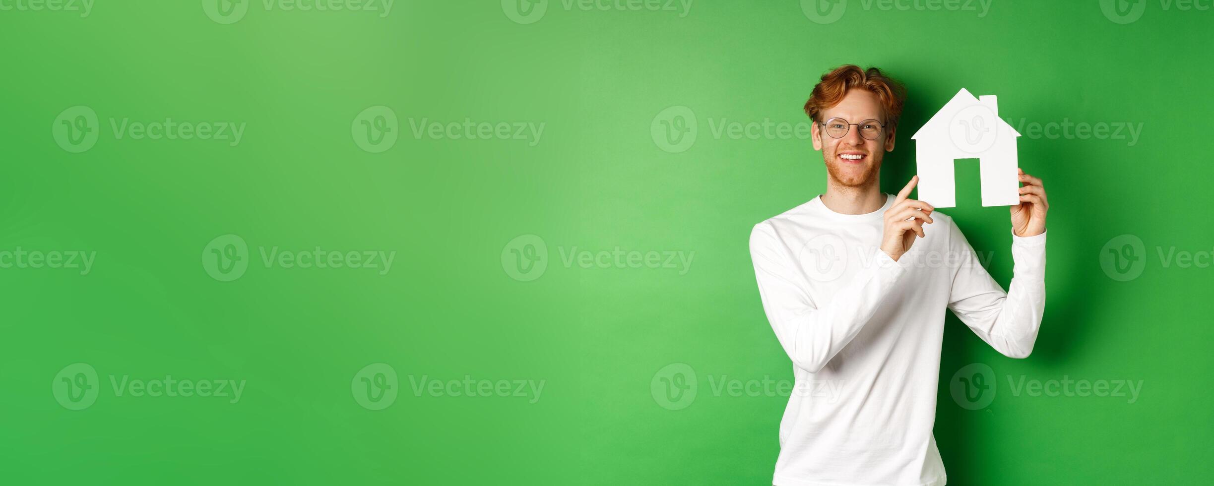 Real estate. Handsome young man with red hair, wearing glasses, showing paper house cutout and smiling, standing against green background photo