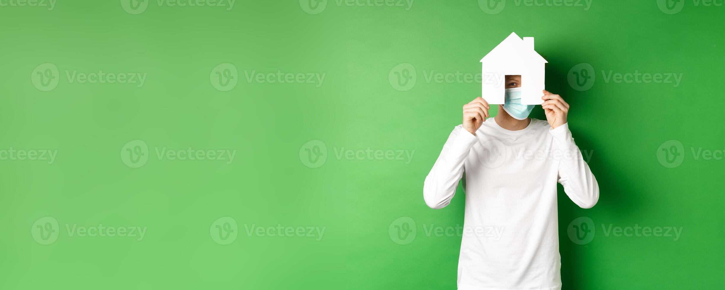Real estate and covid-19 pandemic concept. Funny young man in face mask and white long-sleeve hiding face behind paper house cutout, peeking at camera, green background photo