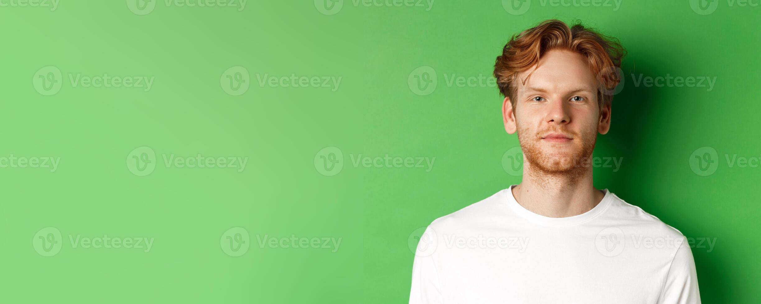 Close-up of young man with red messy hair and beard looking at camera, standing over green background photo