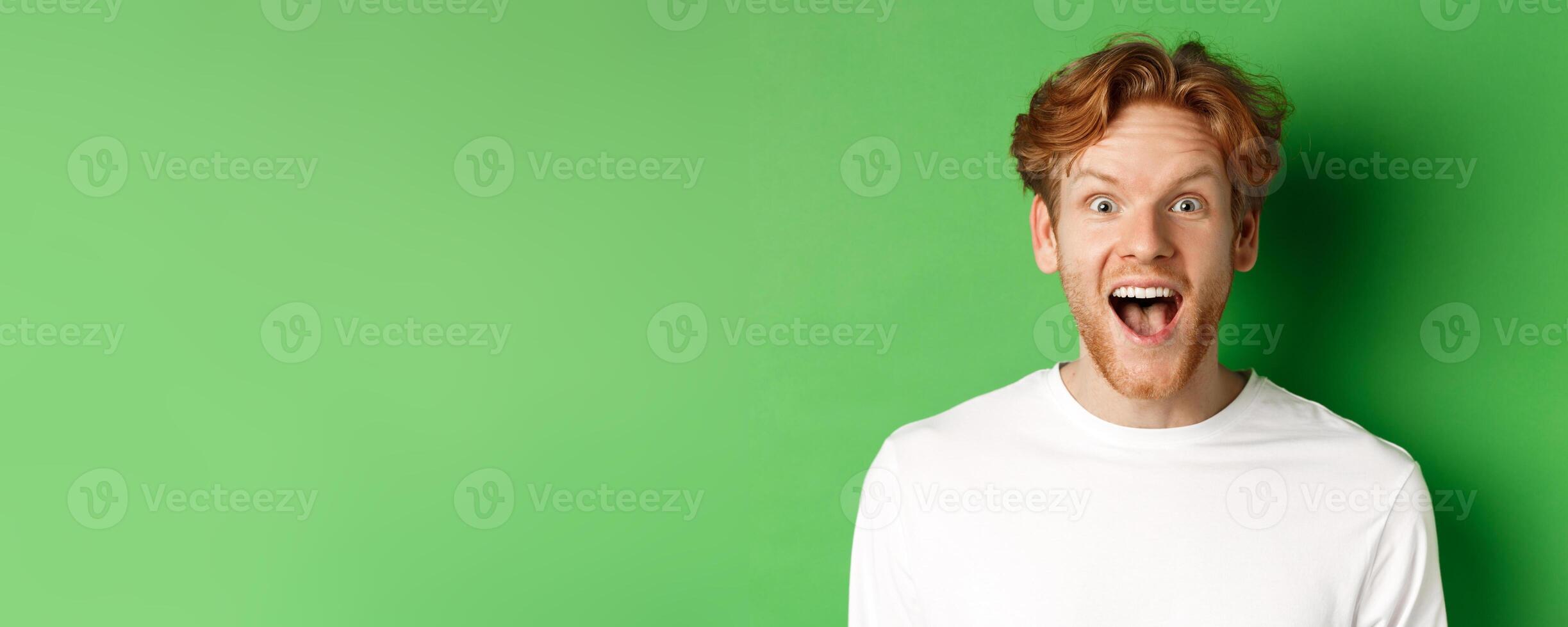 Emotions and fashion concept. Close up of surprised redhead man hear amazing news, staring at camera joyful, standing over green background photo
